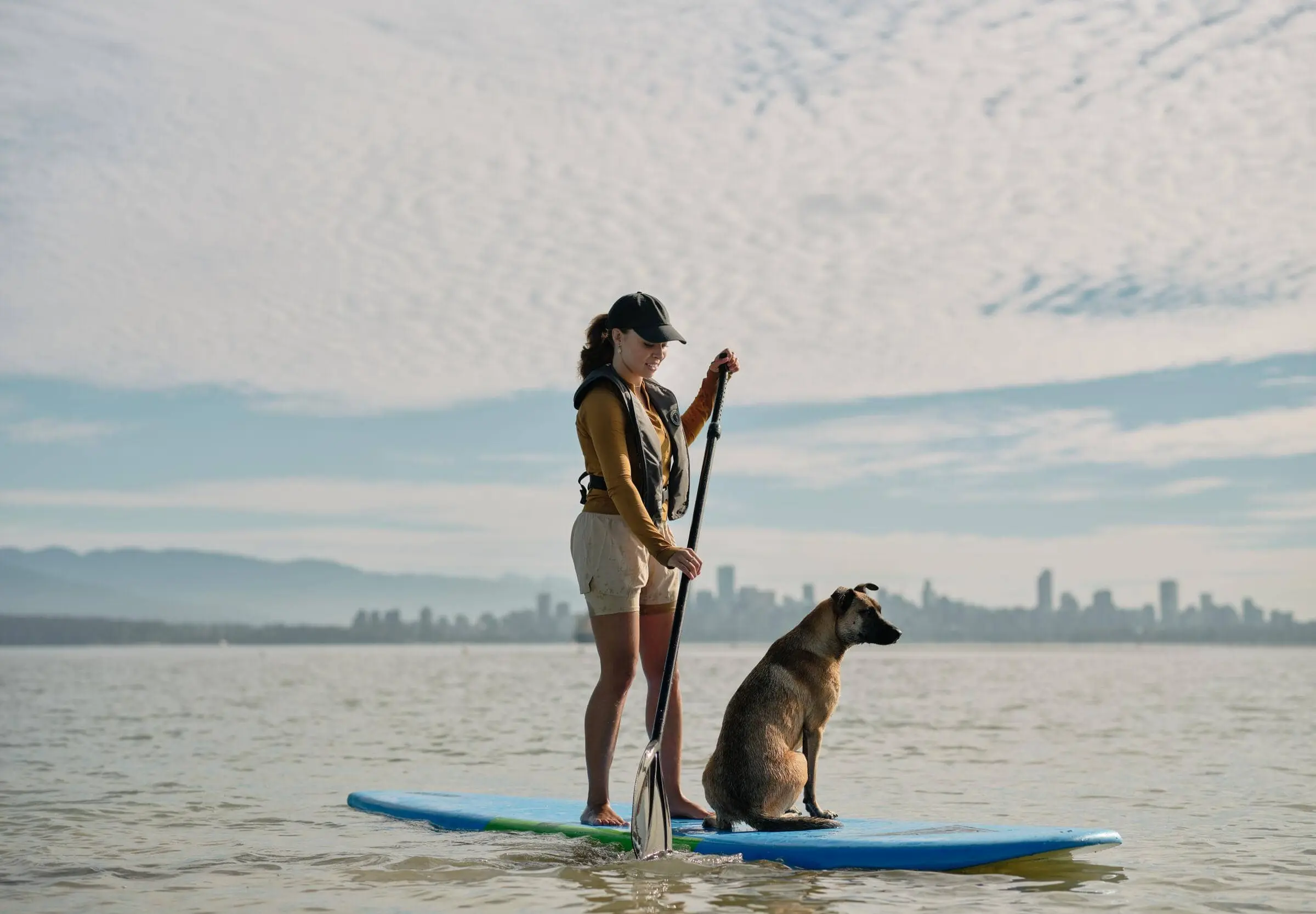Woman in a cap and life jacket paddleboarding on calm water with a dog sitting on the board, city skyline in the background.