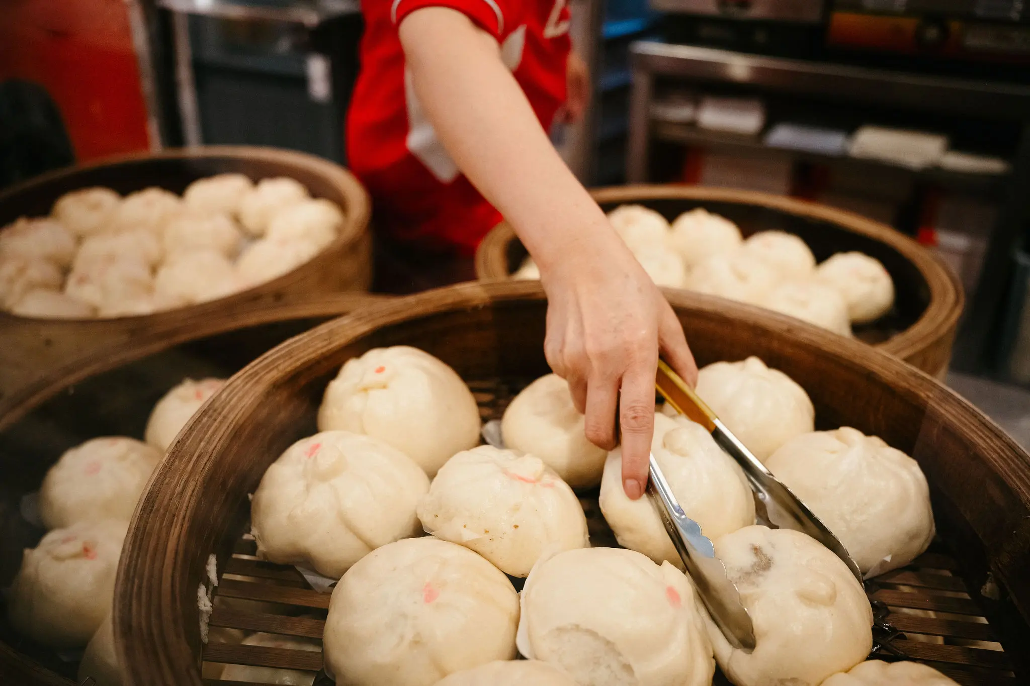 A person using tongs to pick up steamed buns from a bamboo steamer basket filled with buns.