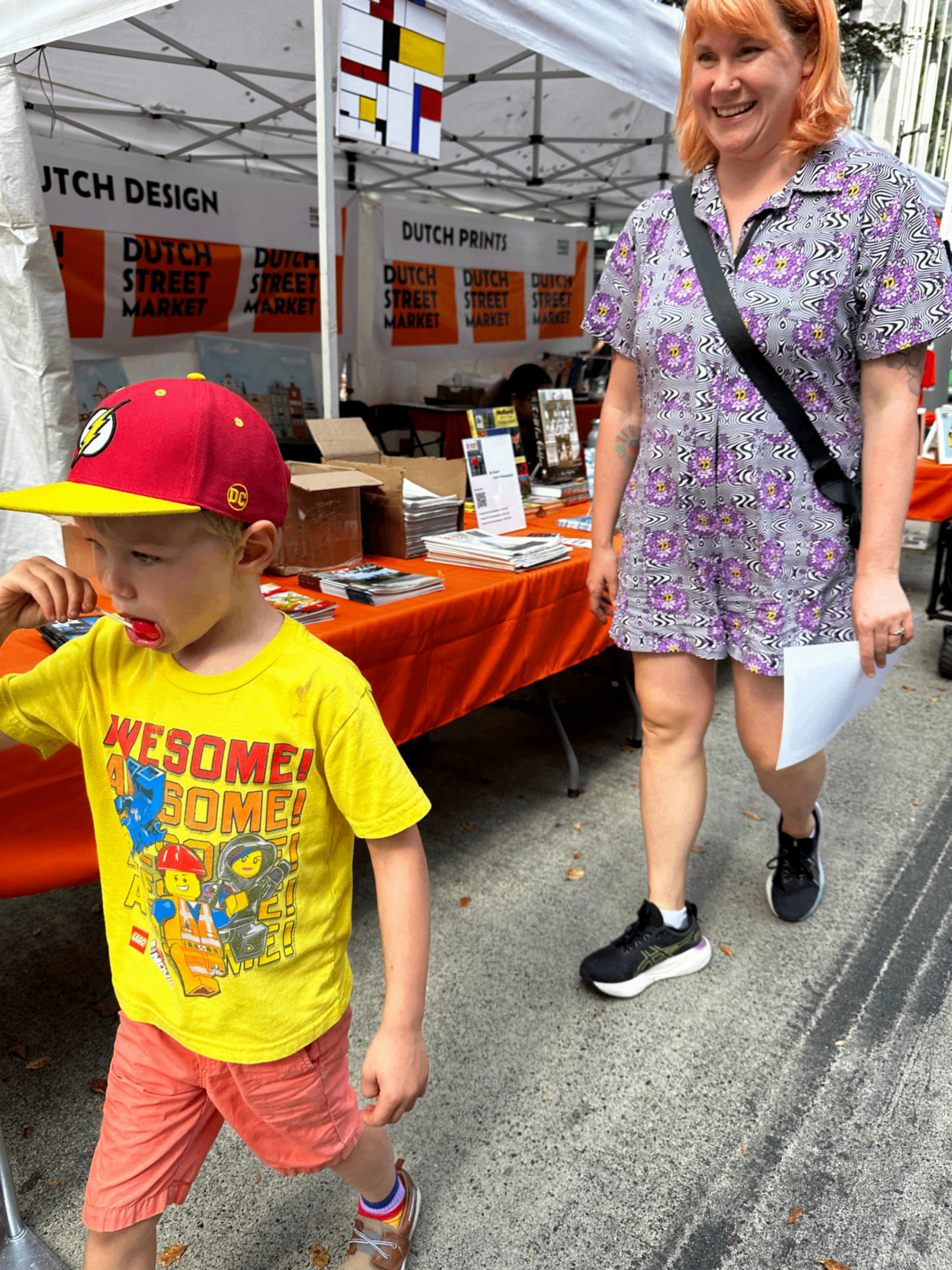 A child in a yellow LEGO t-shirt and red cap walking with a person in a purple floral dress and black sneakers at a Dutch Street Market stall.