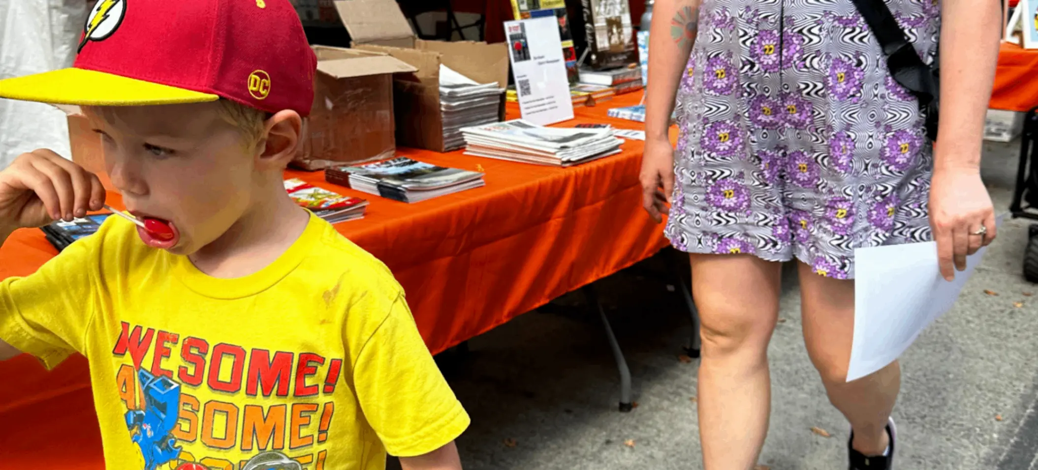 A child in a yellow LEGO t-shirt and red cap walking with a person in a purple floral dress and black sneakers at a Dutch Street Market stall.