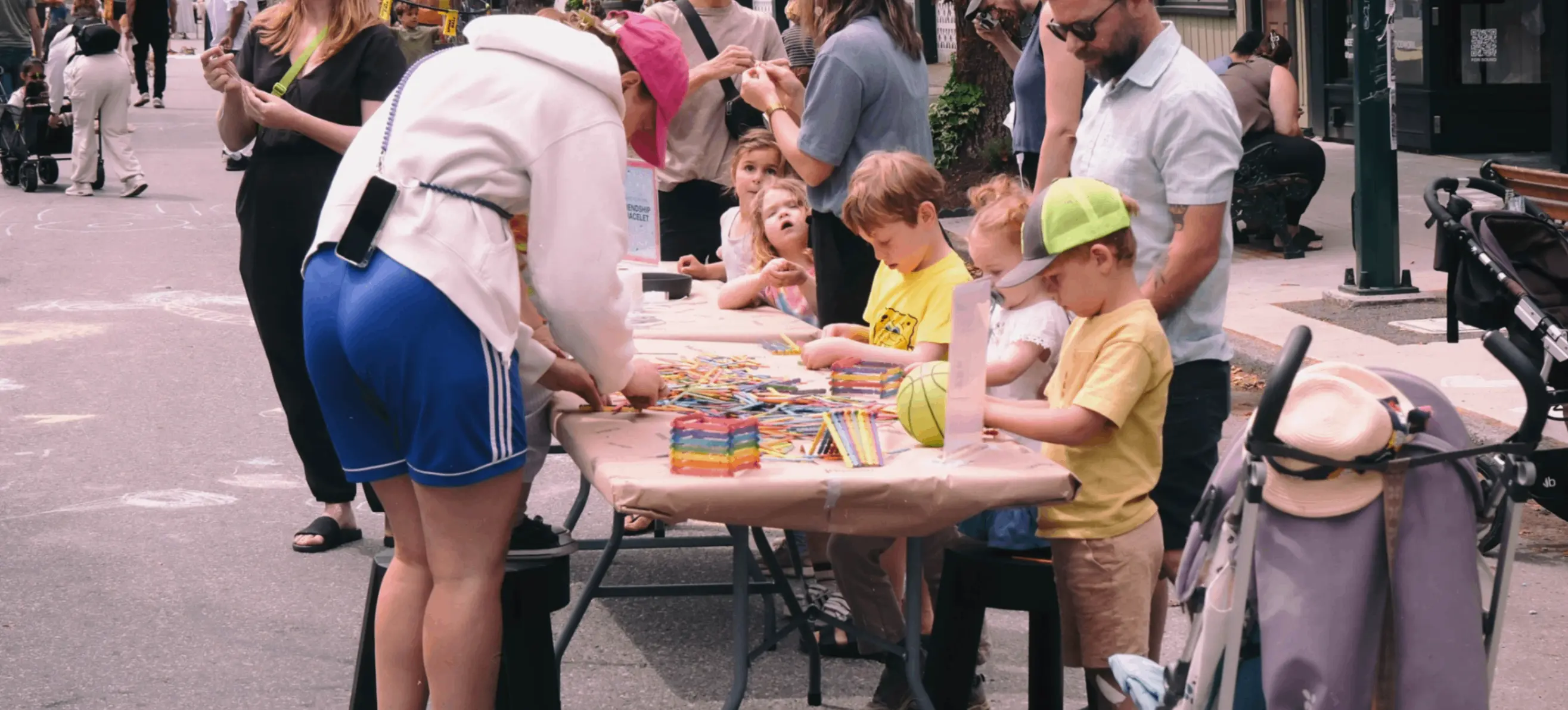 Children and adults gathered around a table outdoors, engaging in arts and crafts with colored pencils and paper.