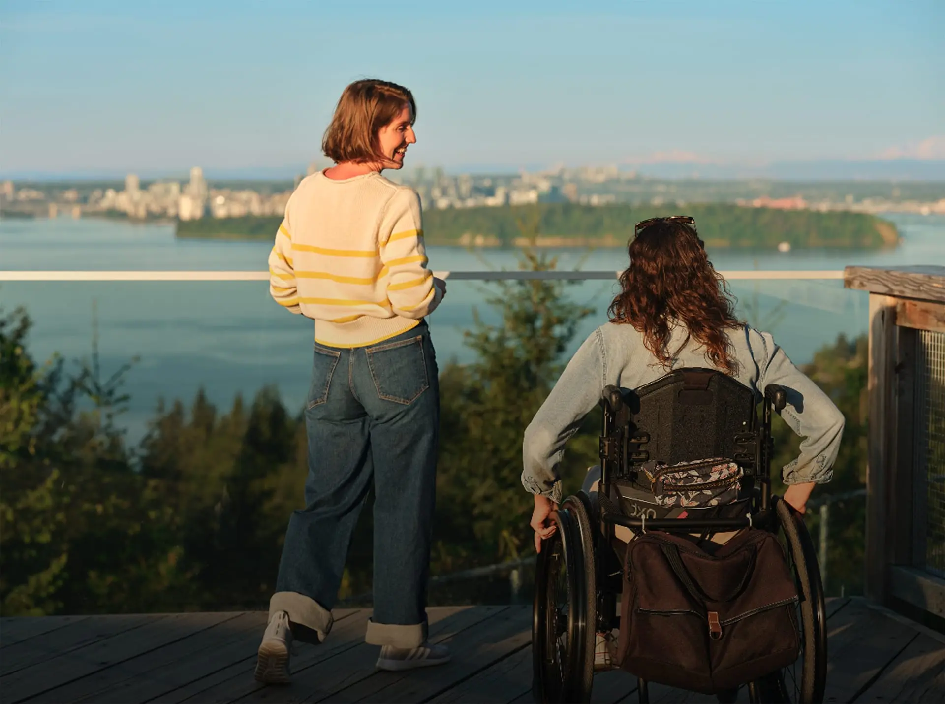 Two women, one standing and one in a wheelchair, overlooking a scenic view of a river and cityscape in the distance.