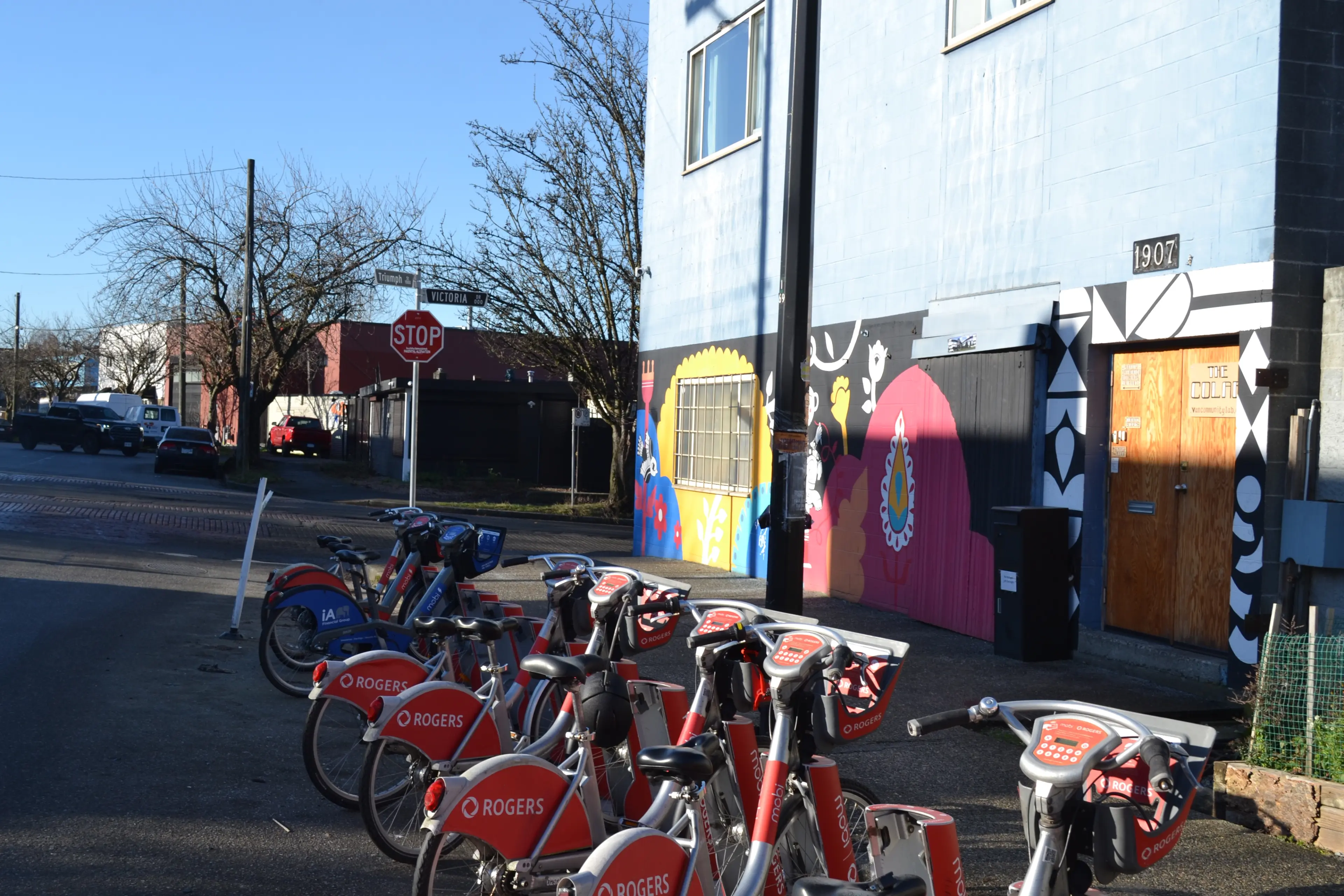 Row of red Rogers rental bikes parked on a street near a colorful mural and a stop sign.