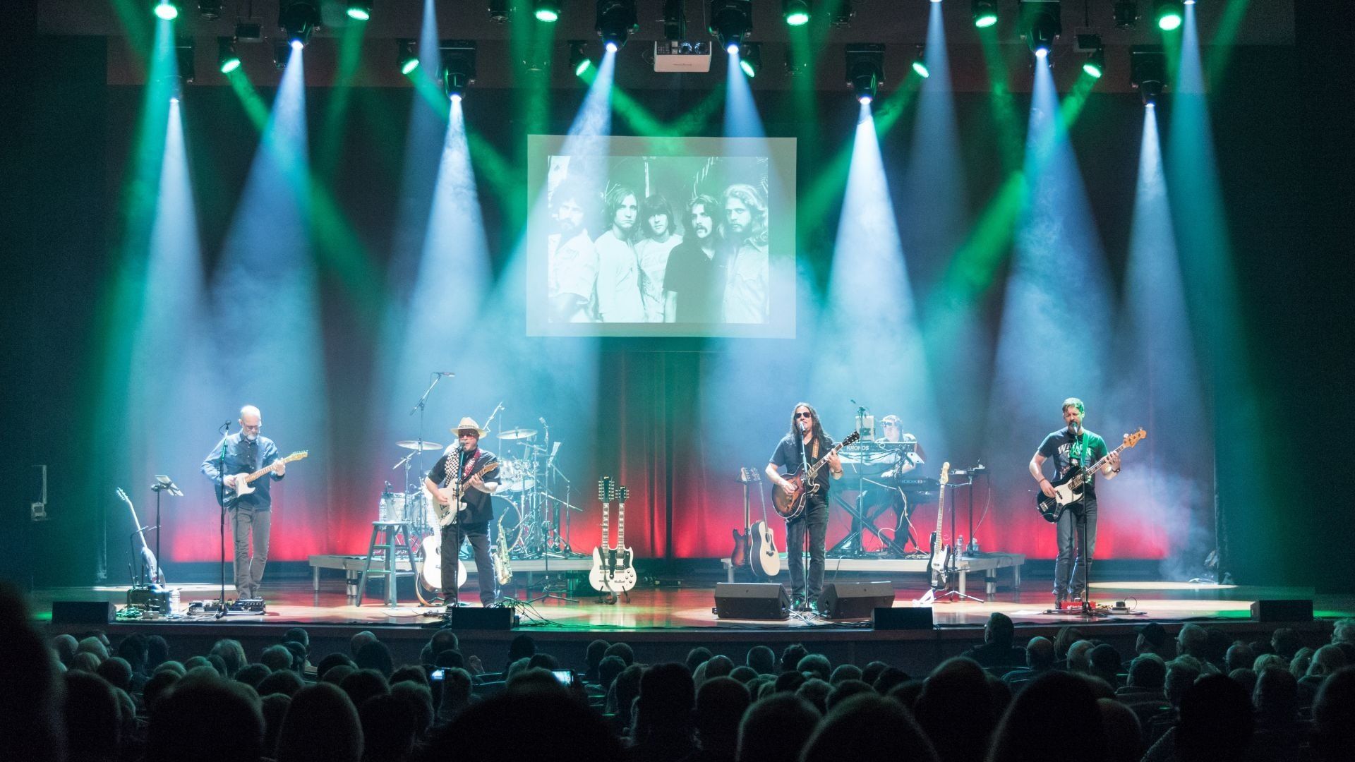 Five musicians performing on stage with guitars and keyboard under green and white spotlights, audience in foreground, black and white band photo projected above.