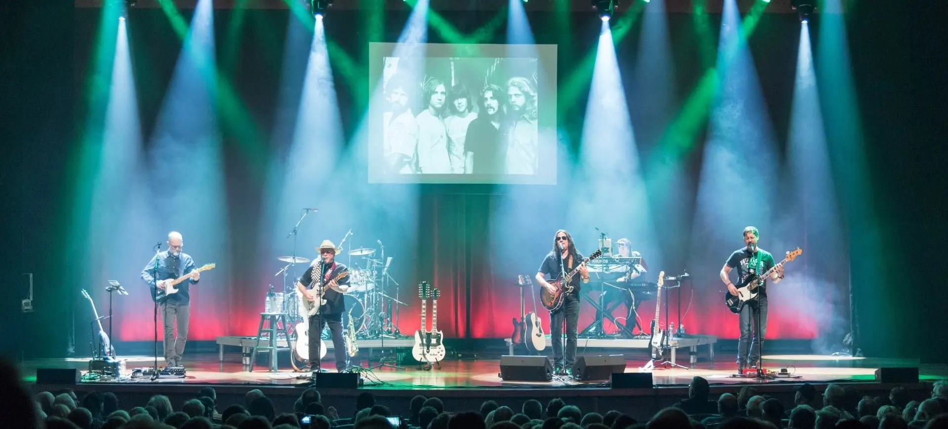 Five musicians performing on stage with guitars and keyboard under green and white spotlights, audience in foreground, black and white band photo projected above.