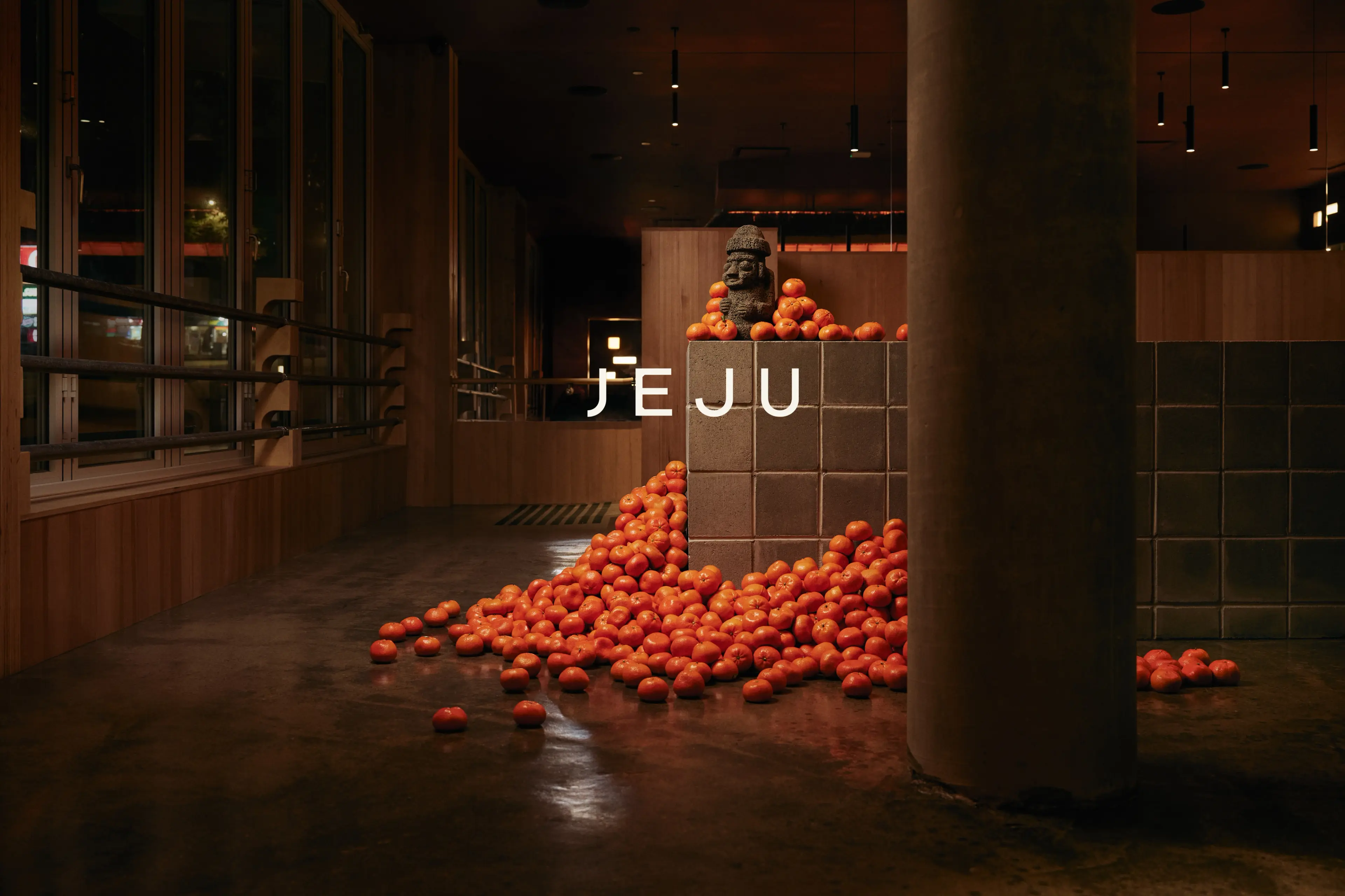 Indoor scene with a pile of orange fruits around a tiled pedestal holding a stone statue, text JEJU overlaid.