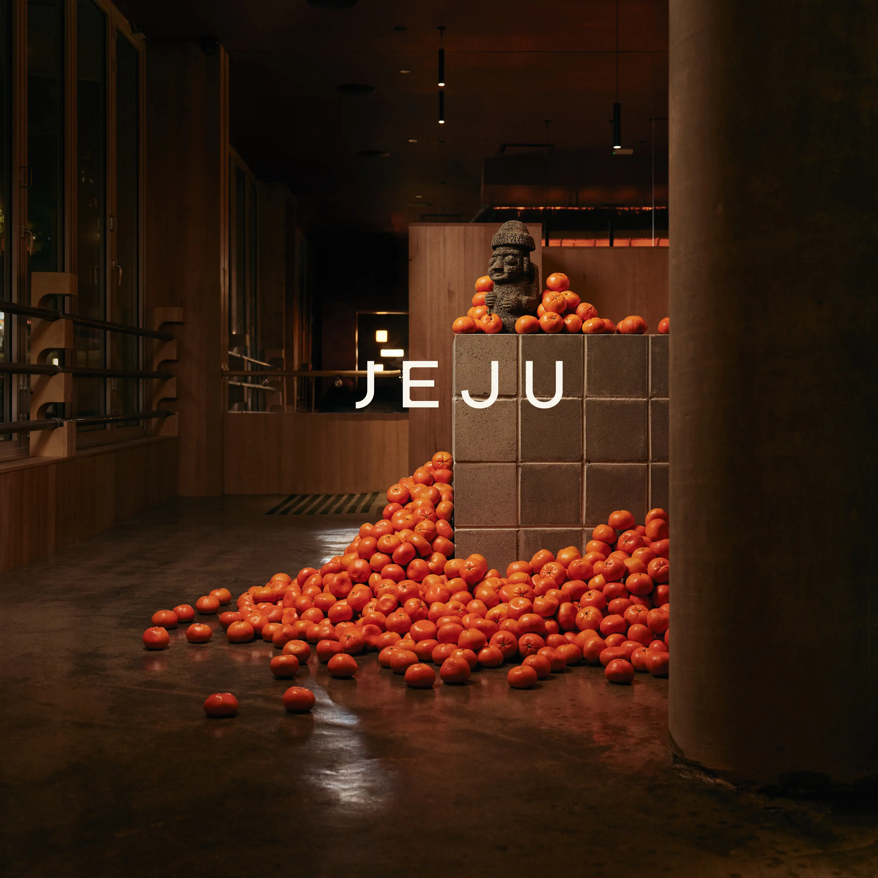 Indoor scene with a pile of orange fruits around a tiled pedestal holding a stone statue, text JEJU overlaid.