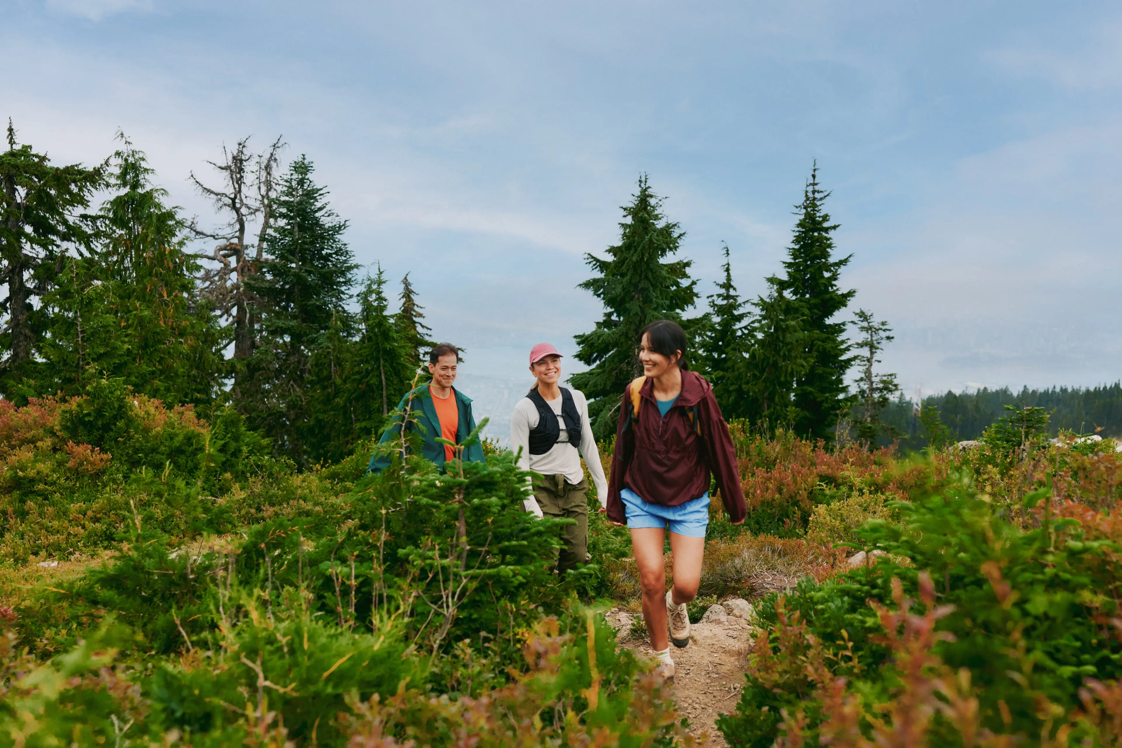 Three people hiking on a trail surrounded by green bushes and tall pine trees under a partly cloudy sky.