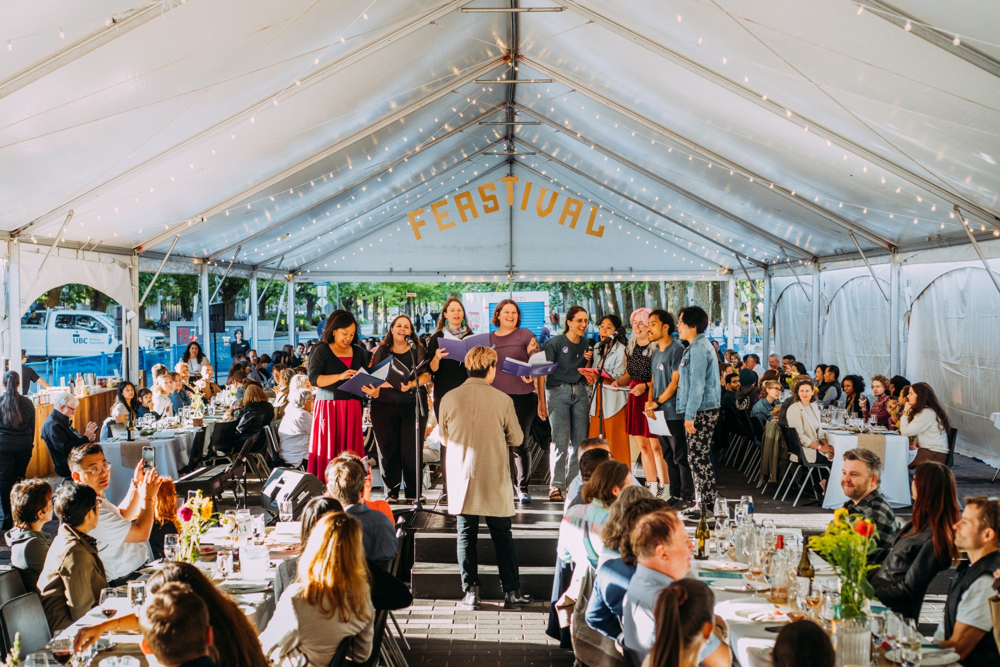 A group of people singing on a stage under a tent with the word FEASTIVAL above, audience seated at tables watching.