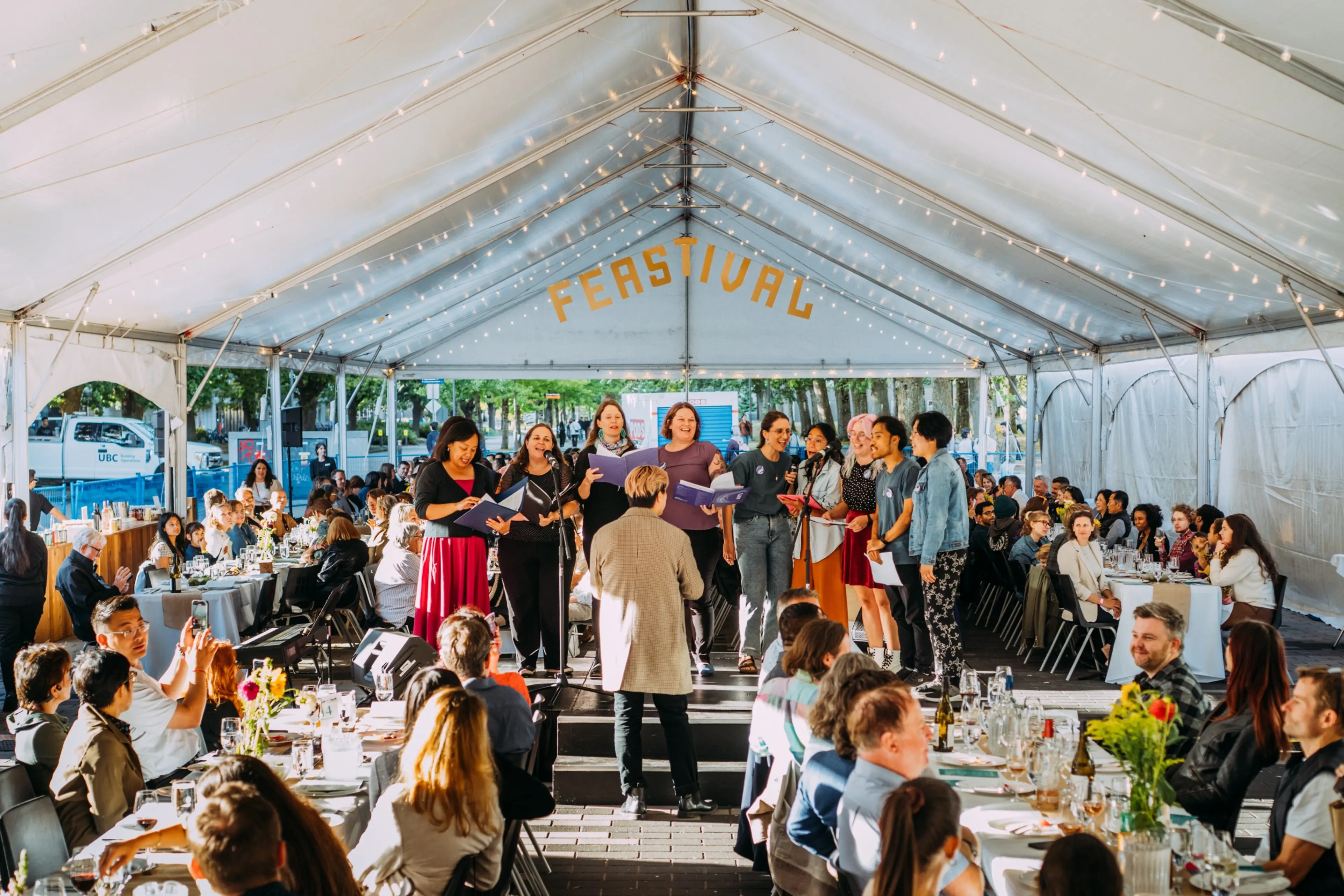 A group of people singing on a stage under a tent with the word FEASTIVAL above, audience seated at tables watching.
