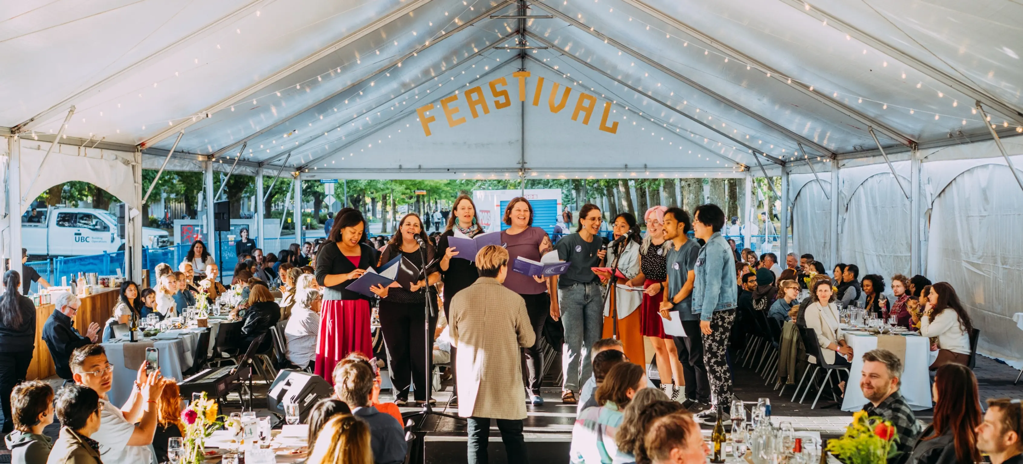A group of people singing on a stage under a tent with the word FEASTIVAL above, audience seated at tables watching.