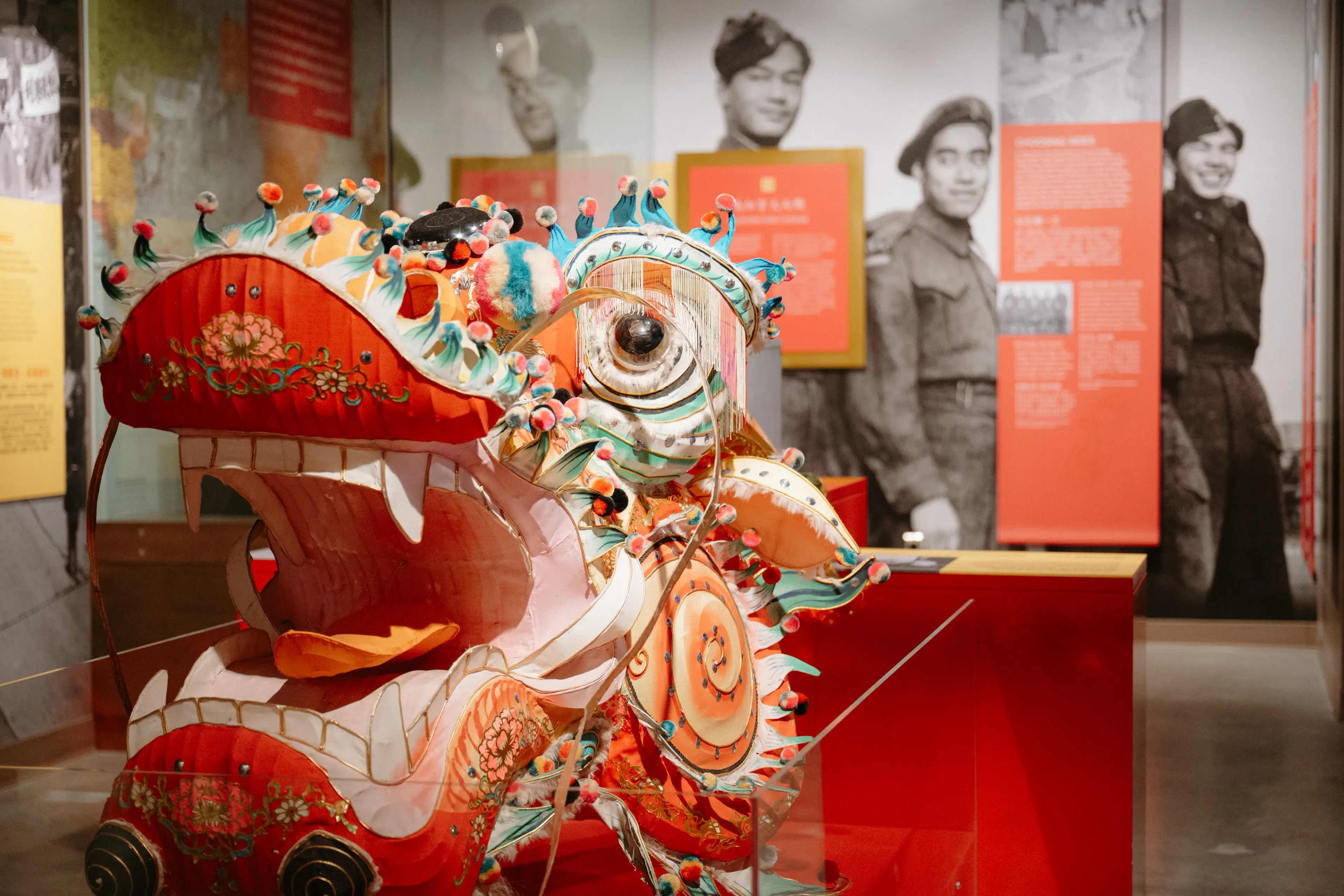 Colorful traditional dragon head sculpture with open mouth displayed in a museum with black and white photos of soldiers in the background.