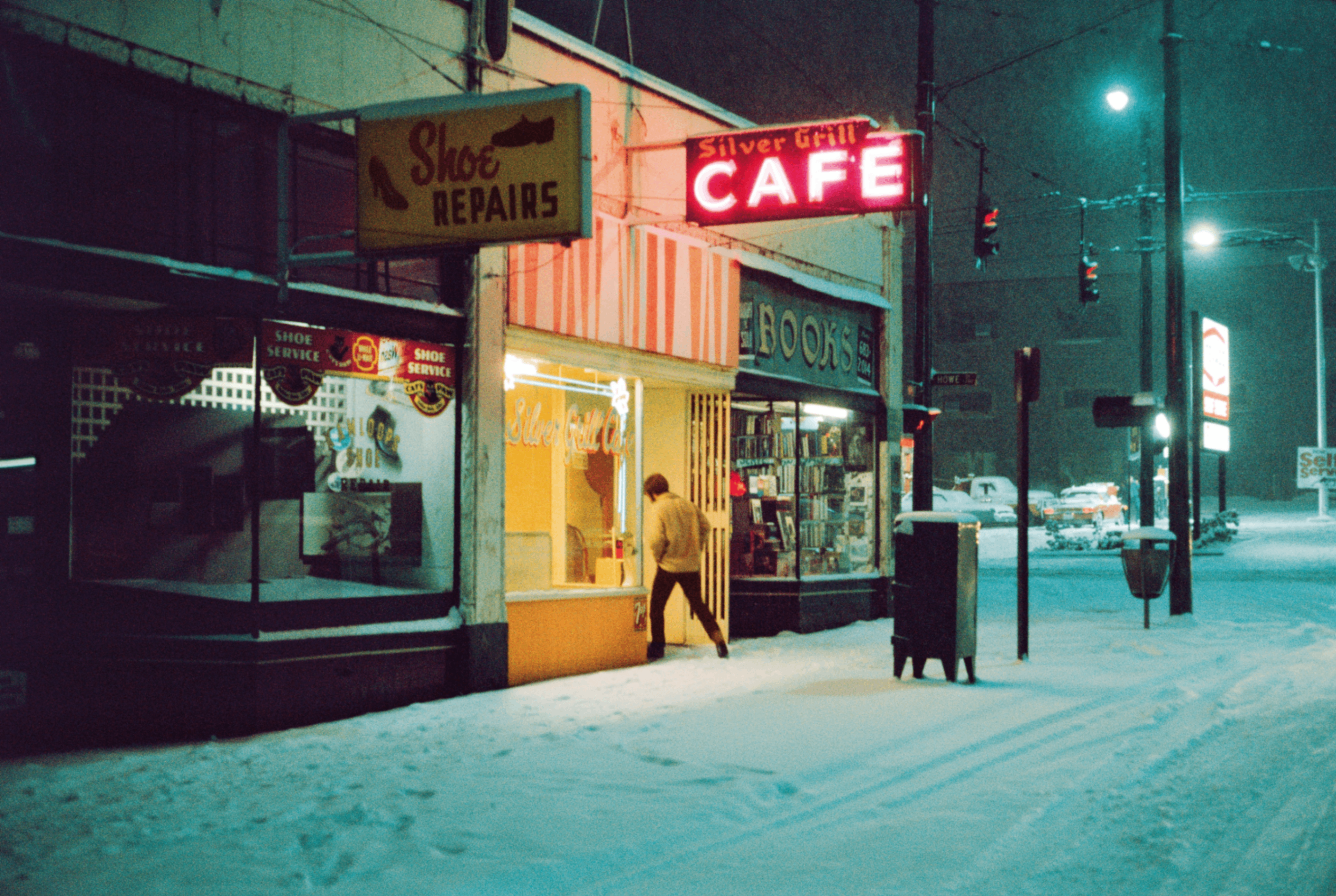 A person entering a brightly lit cafe on a snowy street at night, next to a shoe repair shop and a bookstore.