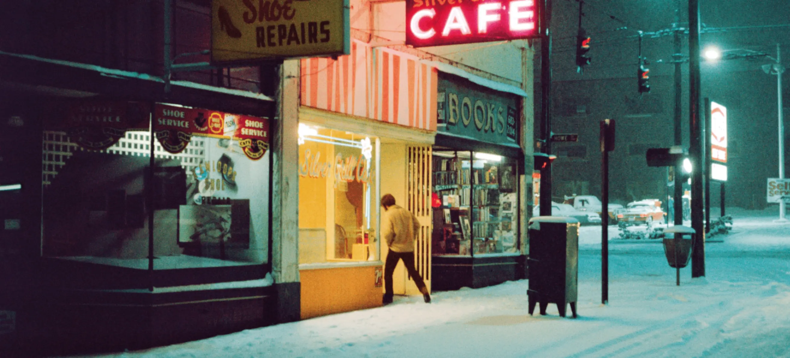A person entering a brightly lit cafe on a snowy street at night, next to a shoe repair shop and a bookstore.