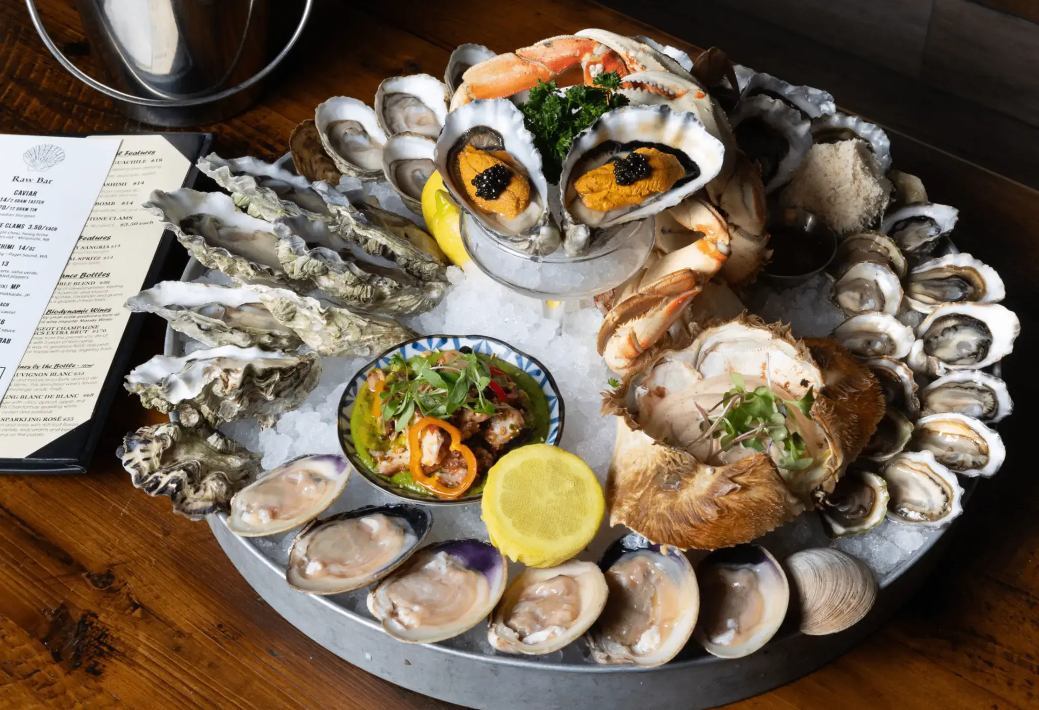 Seafood platter with oysters, crab claws, clams, lemon, and a small dish of garnished seafood on ice with a menu on the side.