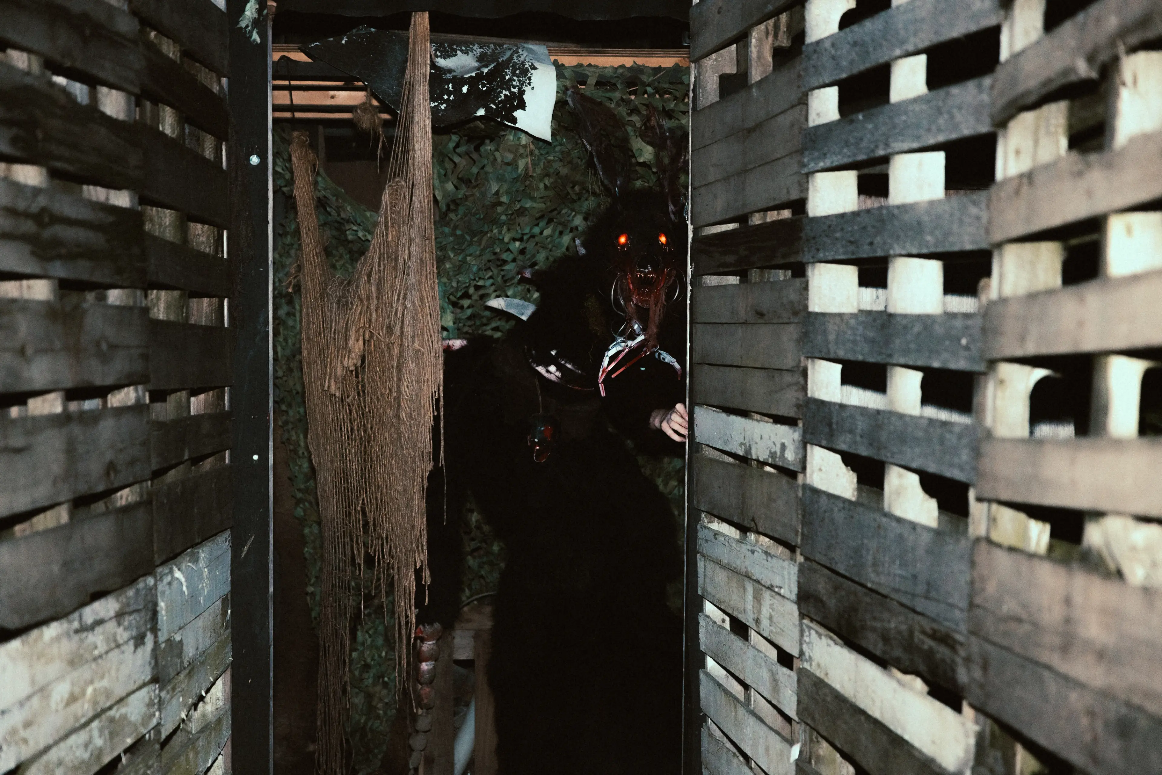 Person wearing a dark costume with glowing red eyes and a mask, standing in a narrow wooden corridor with hanging ropes.