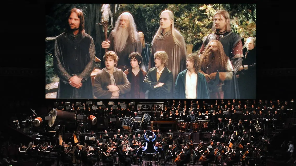Orchestra performing live with conductor in front and a large screen showing characters from a fantasy movie behind them.
