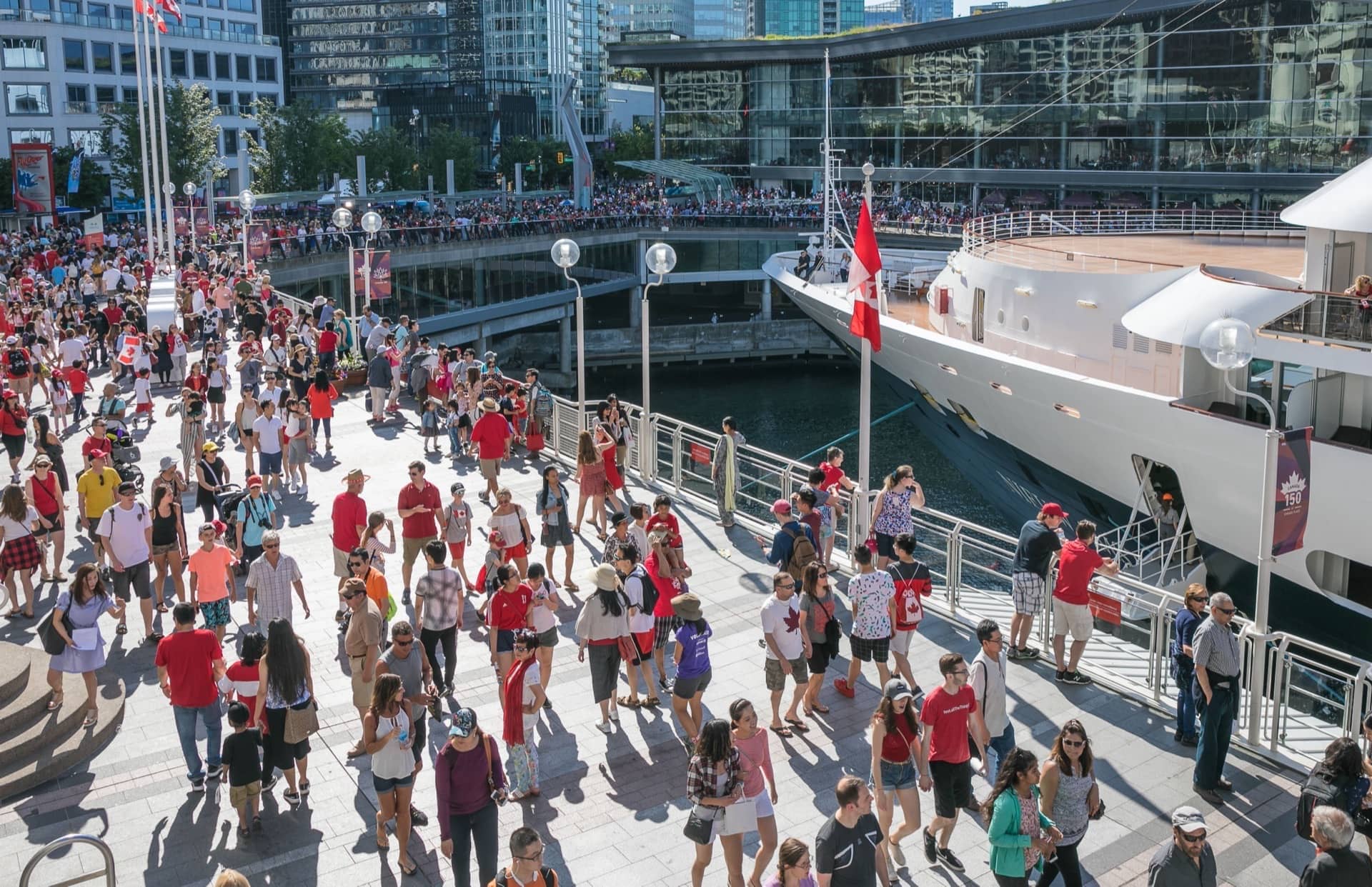 A crowded pedestrian walkway at Canada Place in Vancouver, with people dressed in red and white walking alongside a docked cruise ship and the Vancouver Convention Centre.