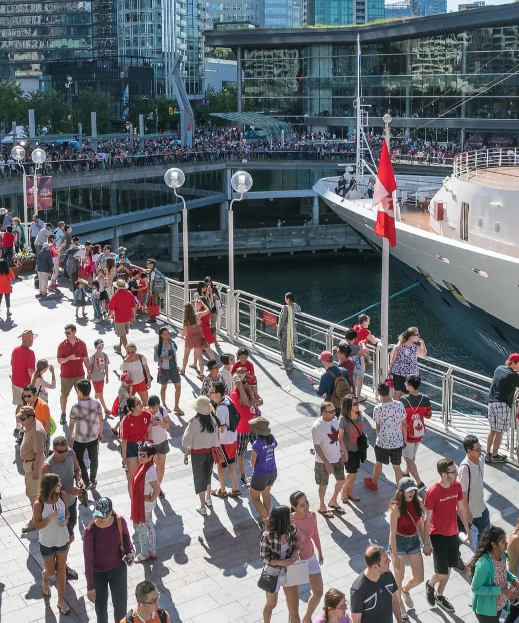 A crowded pedestrian walkway at Canada Place in Vancouver, with people dressed in red and white walking alongside a docked cruise ship and the Vancouver Convention Centre.