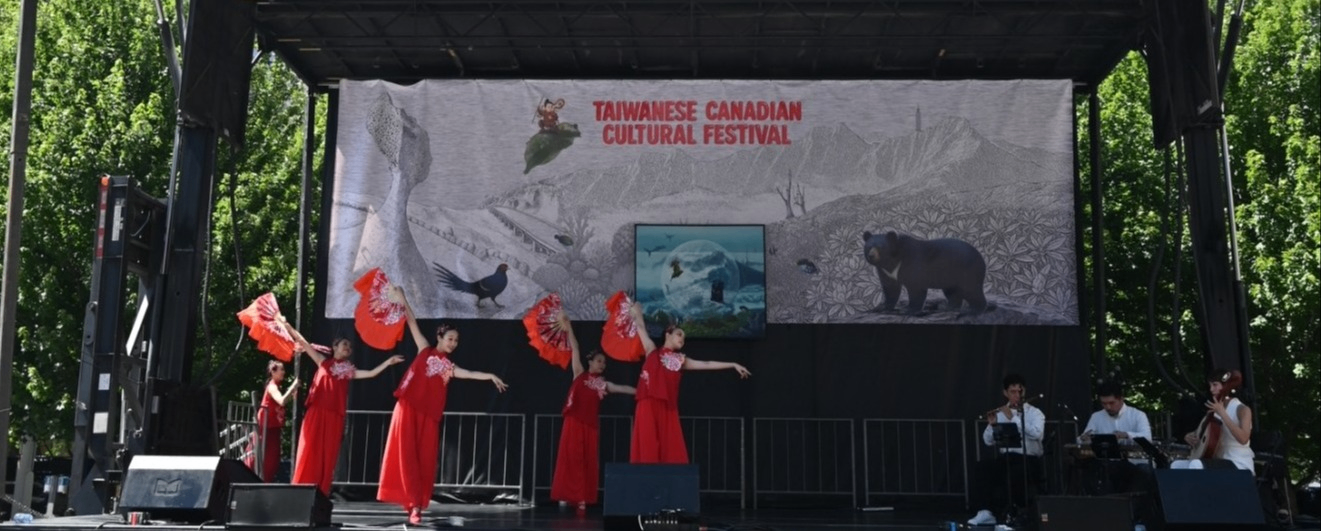 Five dancers in red dresses holding red fans perform on stage at the Taiwanese Canadian Cultural Festival with musicians seated on the right.