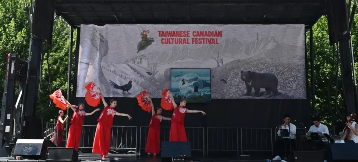 Five dancers in red dresses holding red fans perform on stage at the Taiwanese Canadian Cultural Festival with musicians seated on the right.