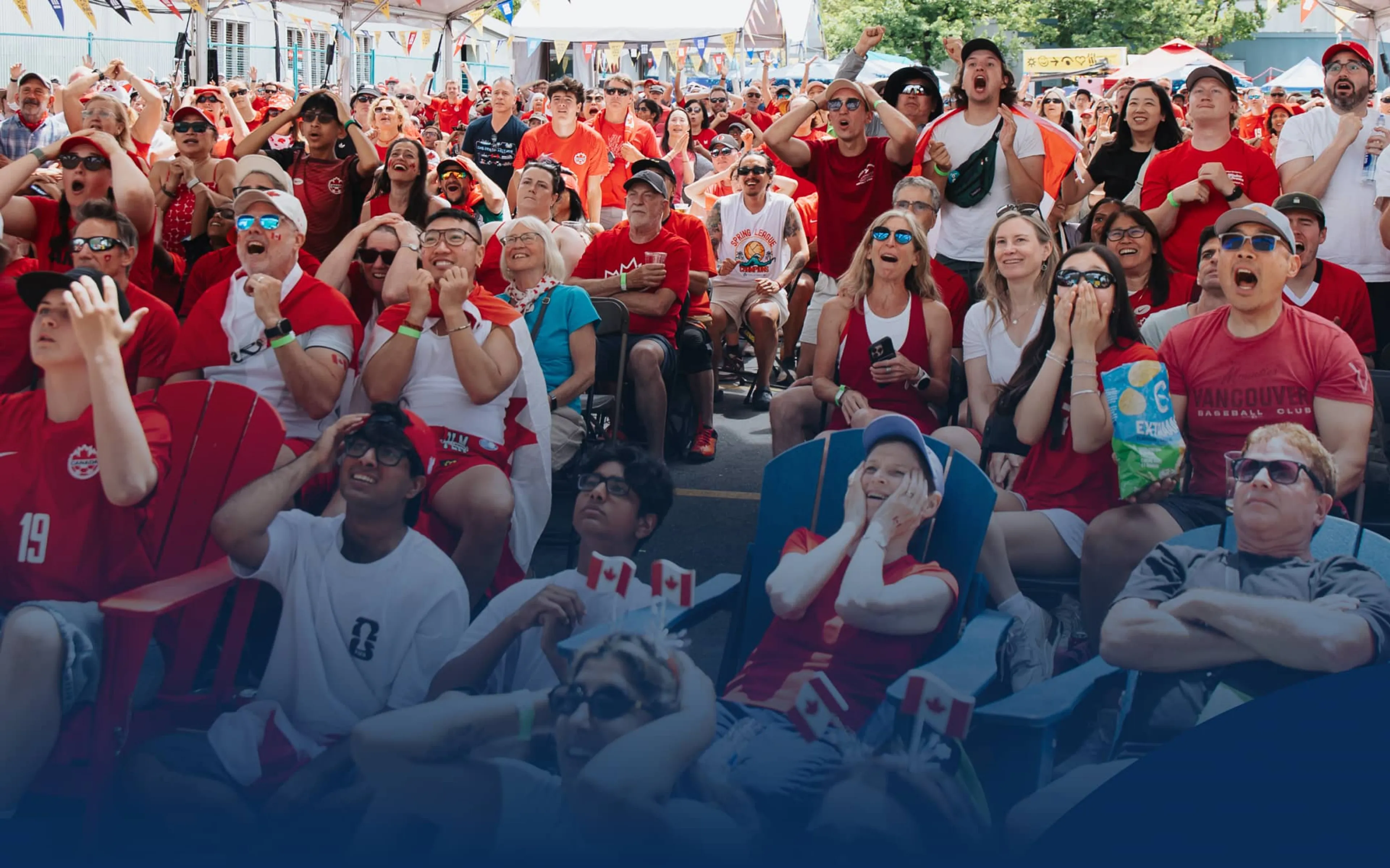 Large crowd of people wearing red and white, watching an event with excited and anxious expressions under a tent with colorful flags.