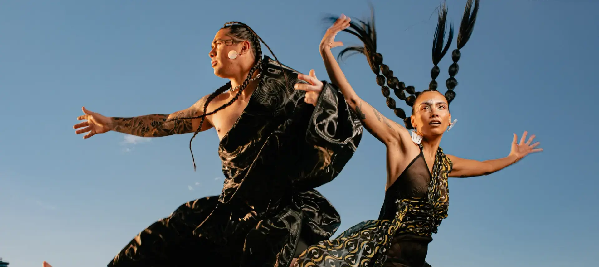 Two people with braided hair wearing flowing black patterned outfits jump energetically against a city skyline and mountain backdrop.