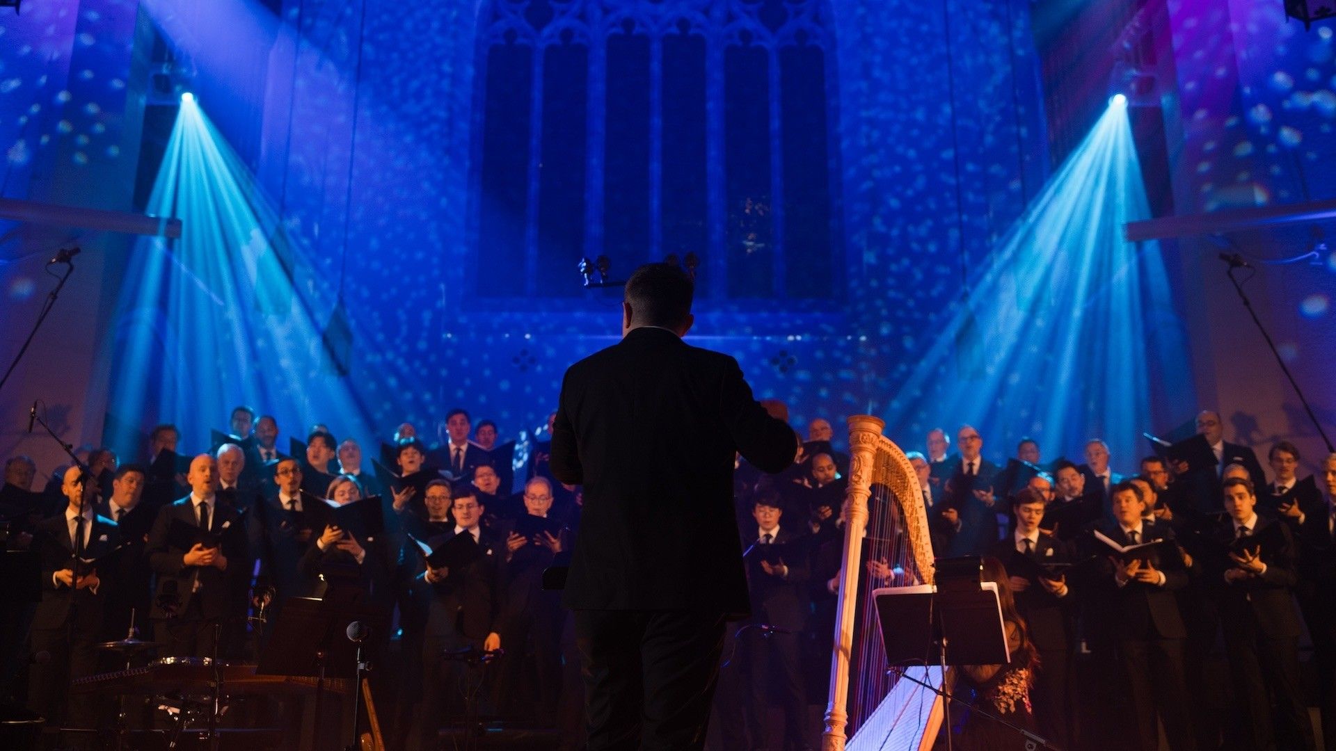 Conductor leading a choir of people in suits with a harp and blue stage lighting in a large hall.