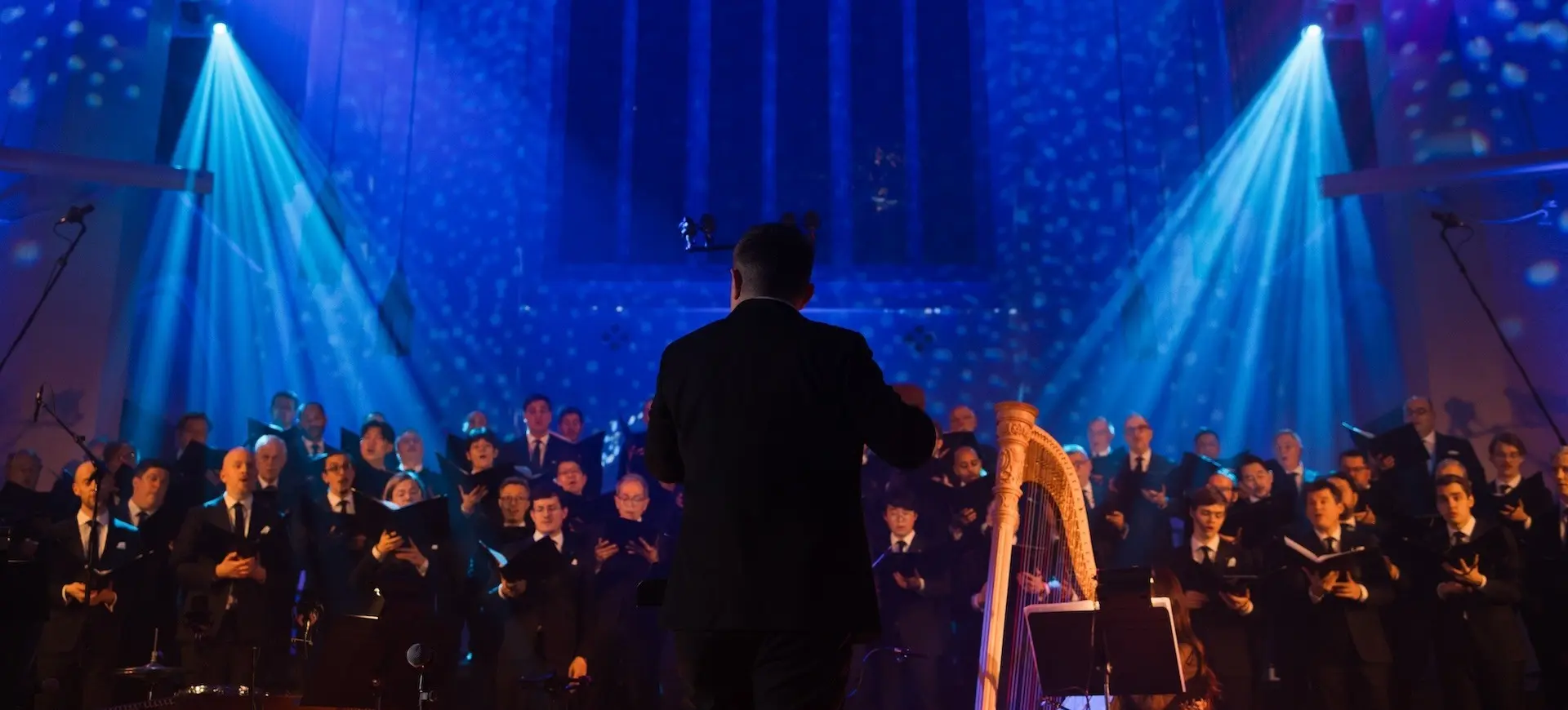 Conductor leading a choir of people in suits with a harp and blue stage lighting in a large hall.