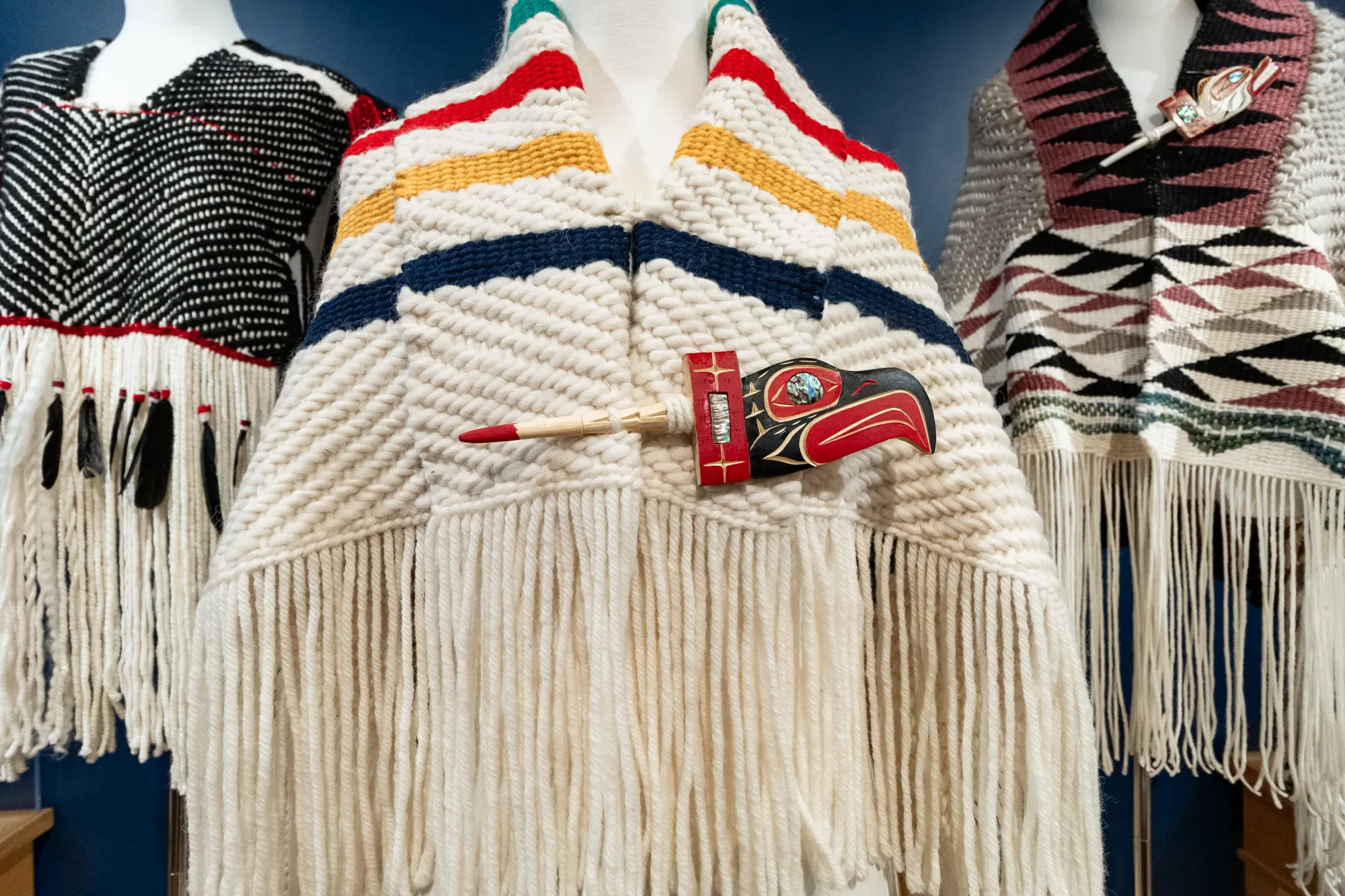 Three woven capes with fringe, each adorned with a carved and painted wooden bird-shaped pin.