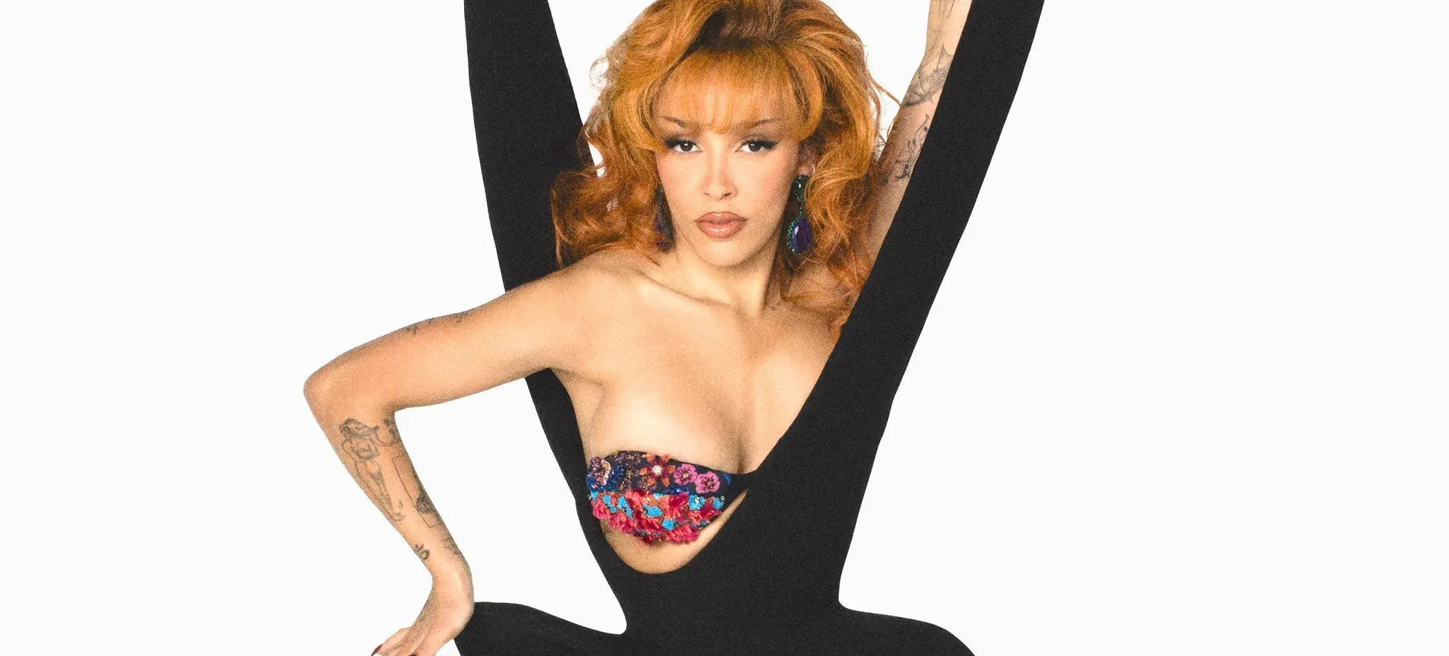 Person with red hair wearing a black outfit with a large V cutout, showing a colorful floral bra, posing with one arm raised and the other on their hip.