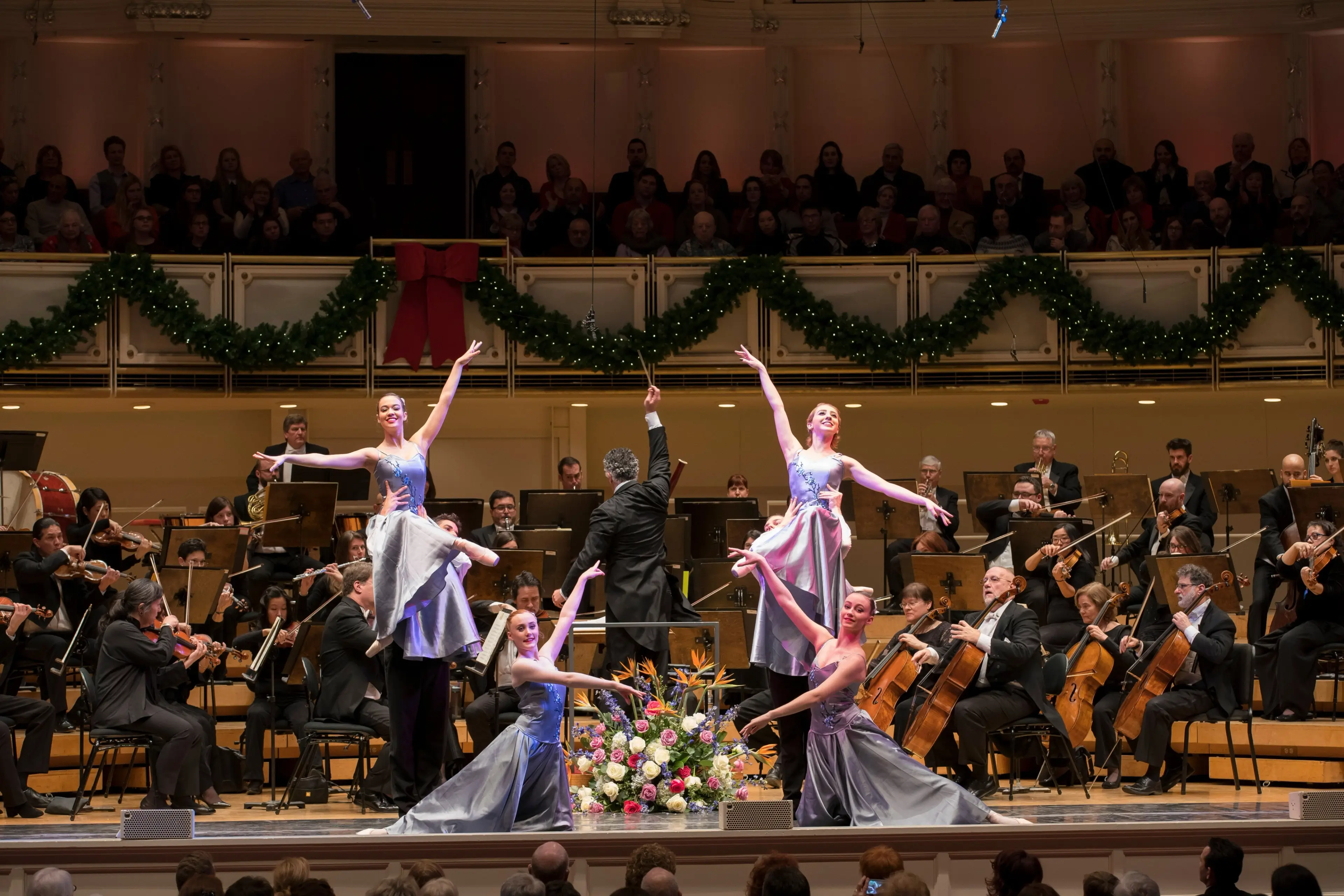 Five ballet dancers in lavender dresses performing on stage with an orchestra and conductor behind them, with holiday garlands and a large floral arrangement.