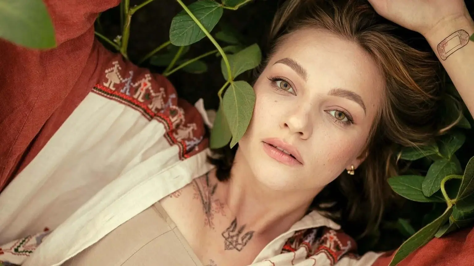 Person with light eyes and light brown hair lying among green leaves, wearing a patterned red and white garment, with tattoos on neck and wrist.