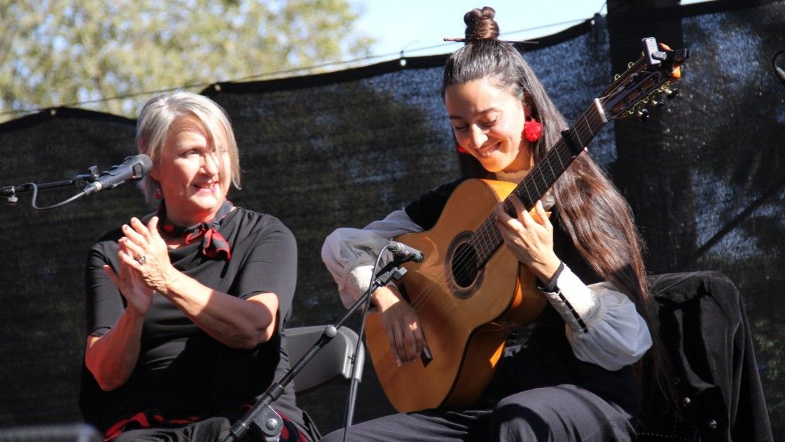 Two people on stage, one clapping and the other playing an acoustic guitar, both smiling.