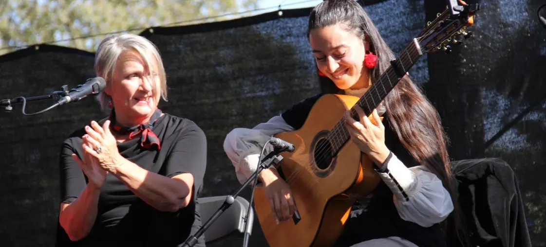 Two people on stage, one clapping and the other playing an acoustic guitar, both smiling.