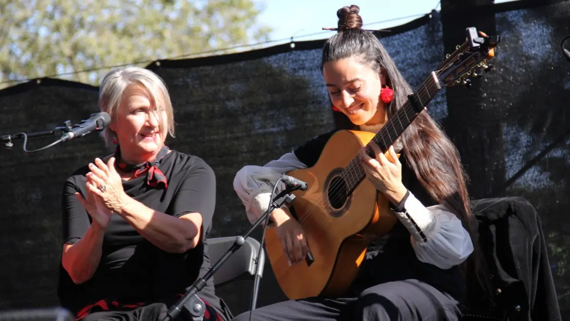 Two people on stage, one clapping and the other playing an acoustic guitar, both smiling.