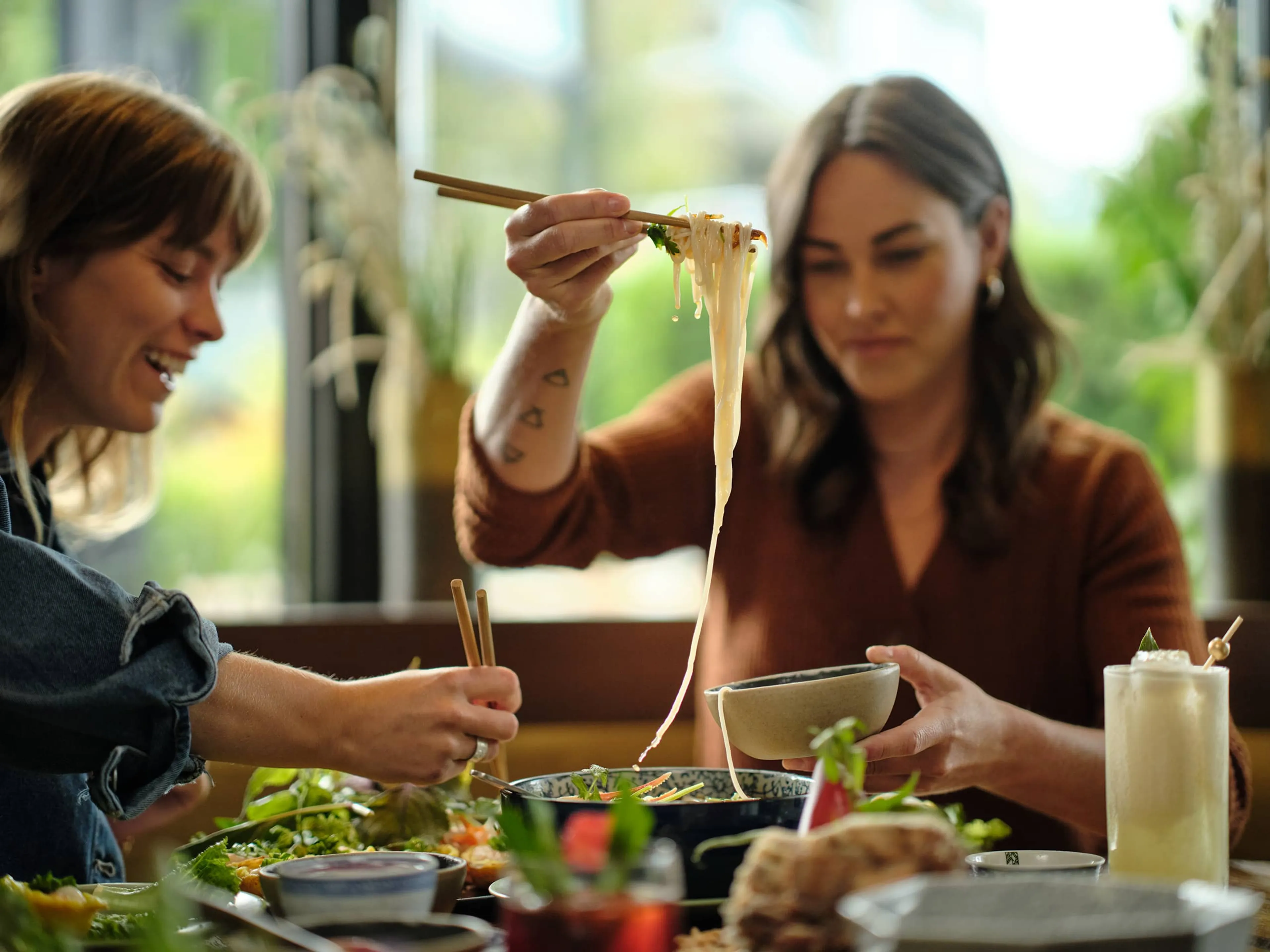 Two women using chopsticks to eat noodles at a table with various dishes and a drink.