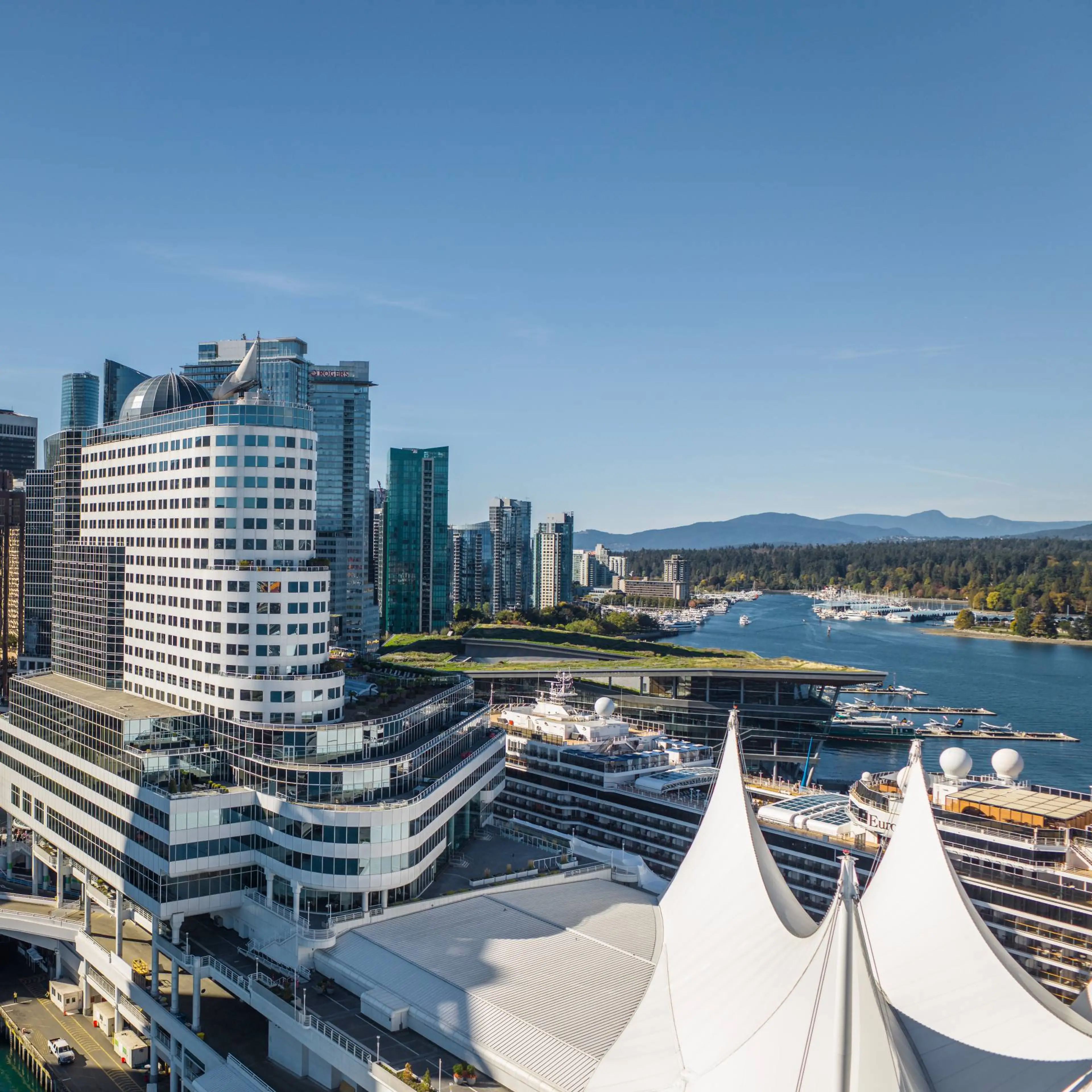 City skyline with modern buildings, waterfront, cruise ships, and white tent-like roof structures under clear blue sky.