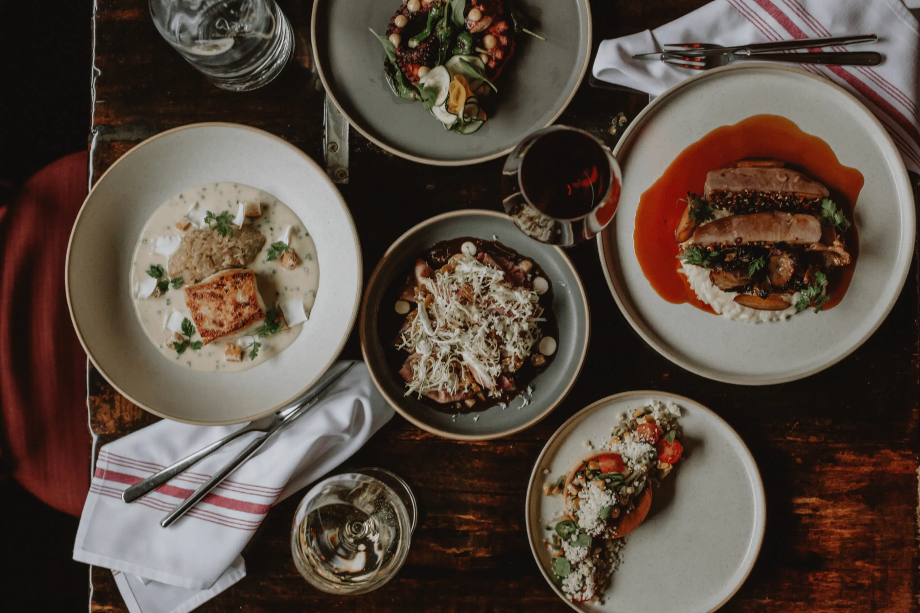 Five plates of gourmet dishes with various ingredients and two glasses of wine on a wooden table with white napkins and cutlery.