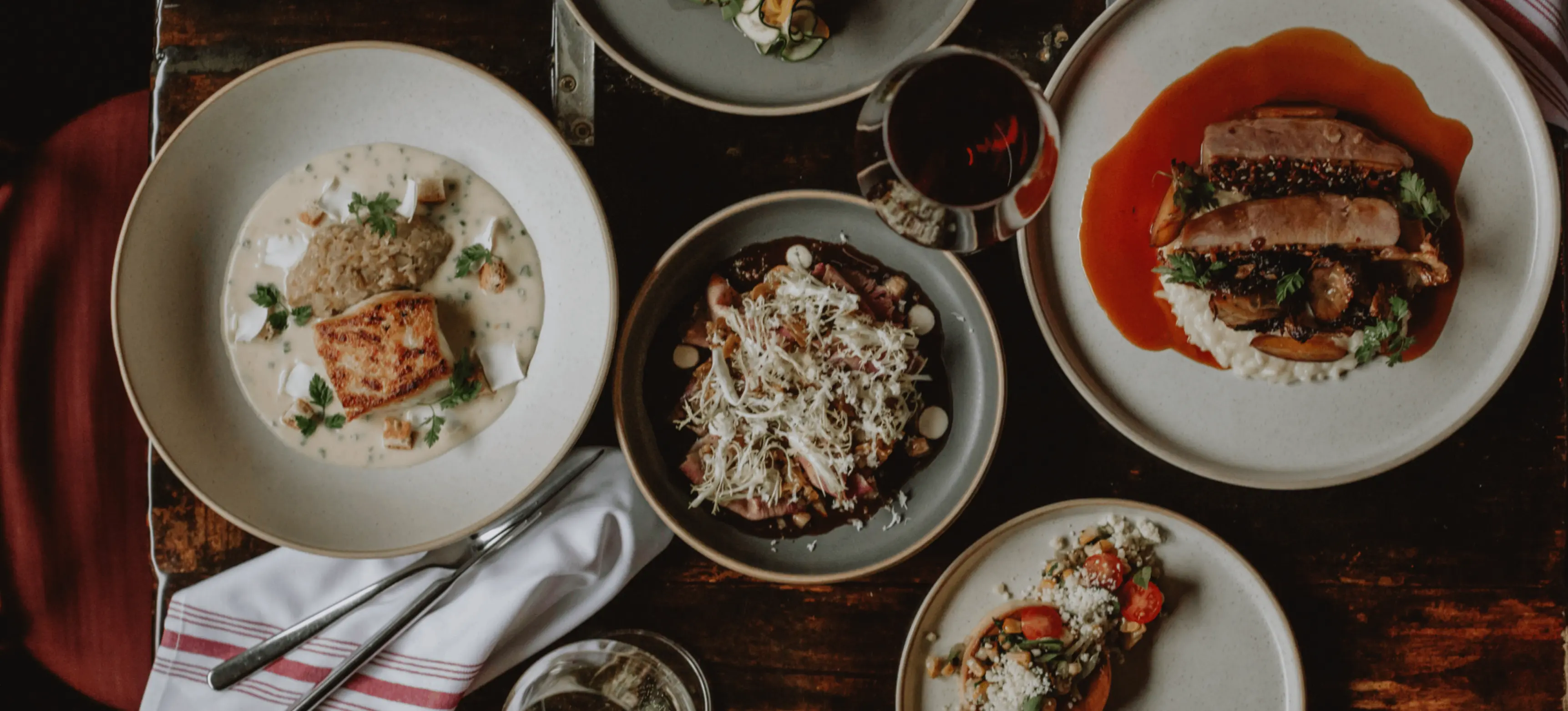 Five plates of gourmet dishes with various ingredients and two glasses of wine on a wooden table with white napkins and cutlery.