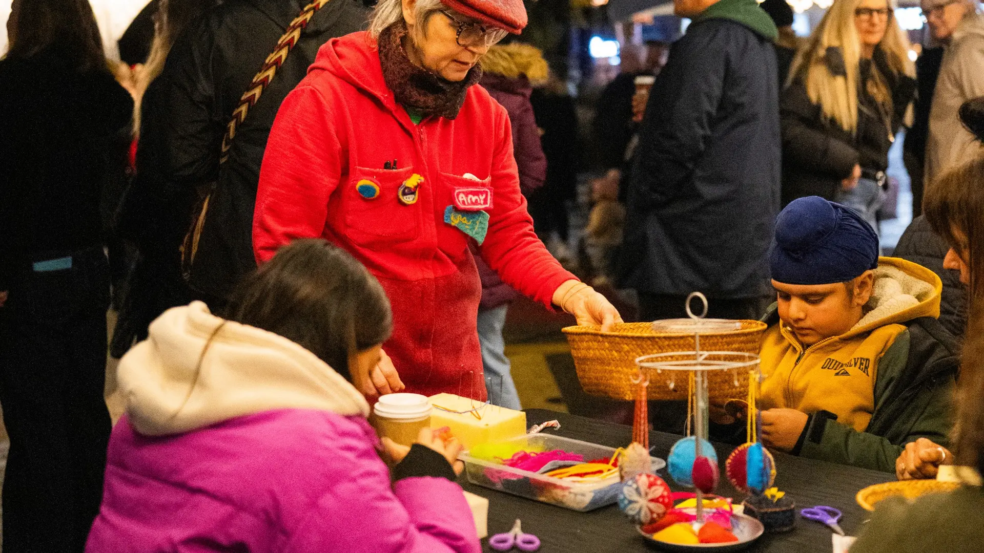 A person in a red jacket and hat assists two children at a craft table with colorful materials and hanging decorations.