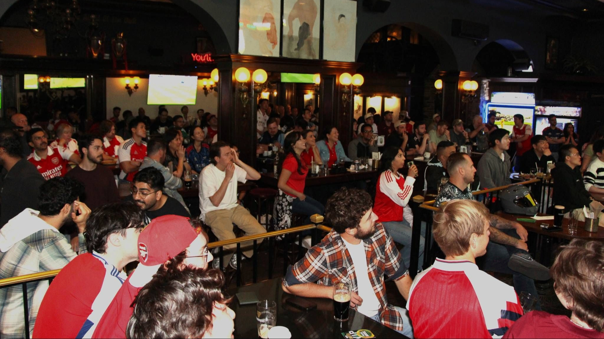 A large group of people seated indoors watching multiple screens, some wearing sports jerseys, in a dimly lit bar or pub.