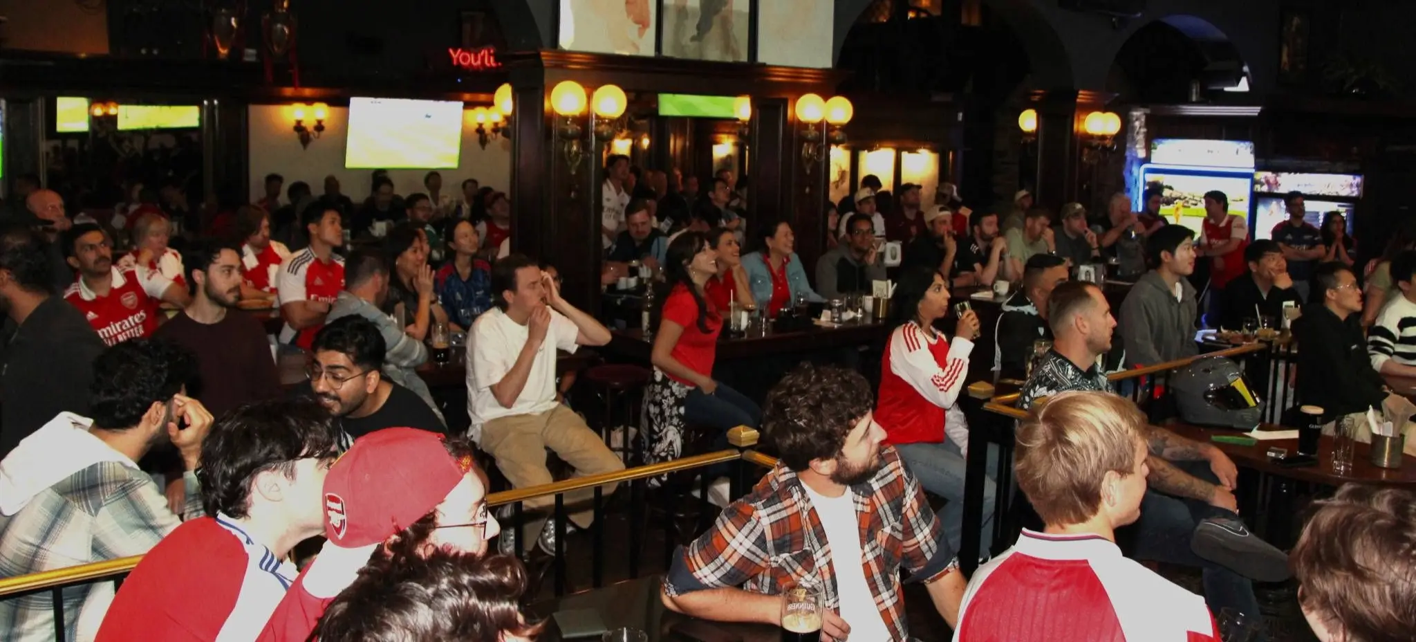 A large group of people seated indoors watching multiple screens, some wearing sports jerseys, in a dimly lit bar or pub.