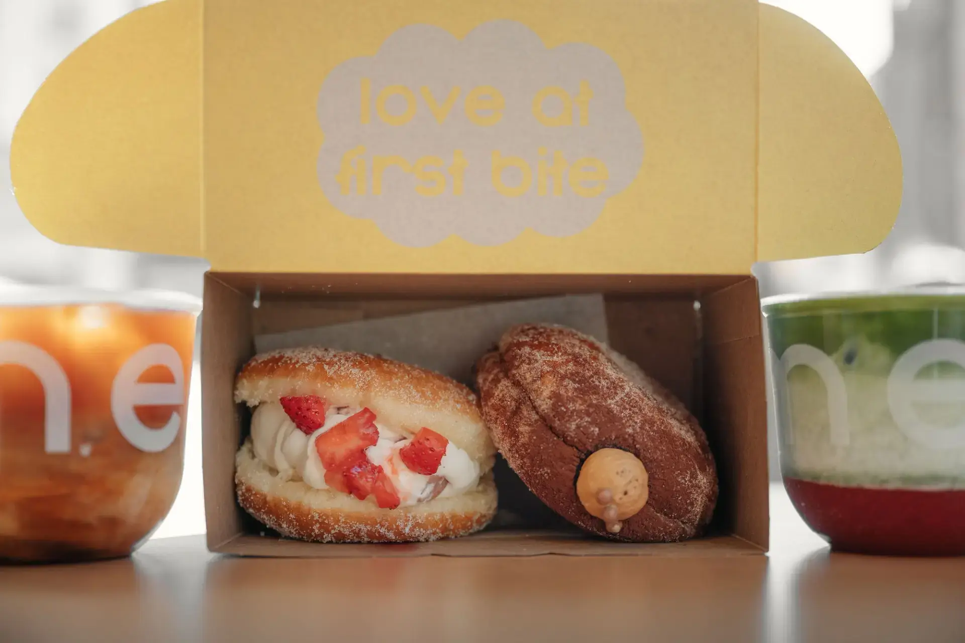 Two cream-filled pastries with strawberries and caramel in a box labeled love at first bite, flanked by two colorful cups.