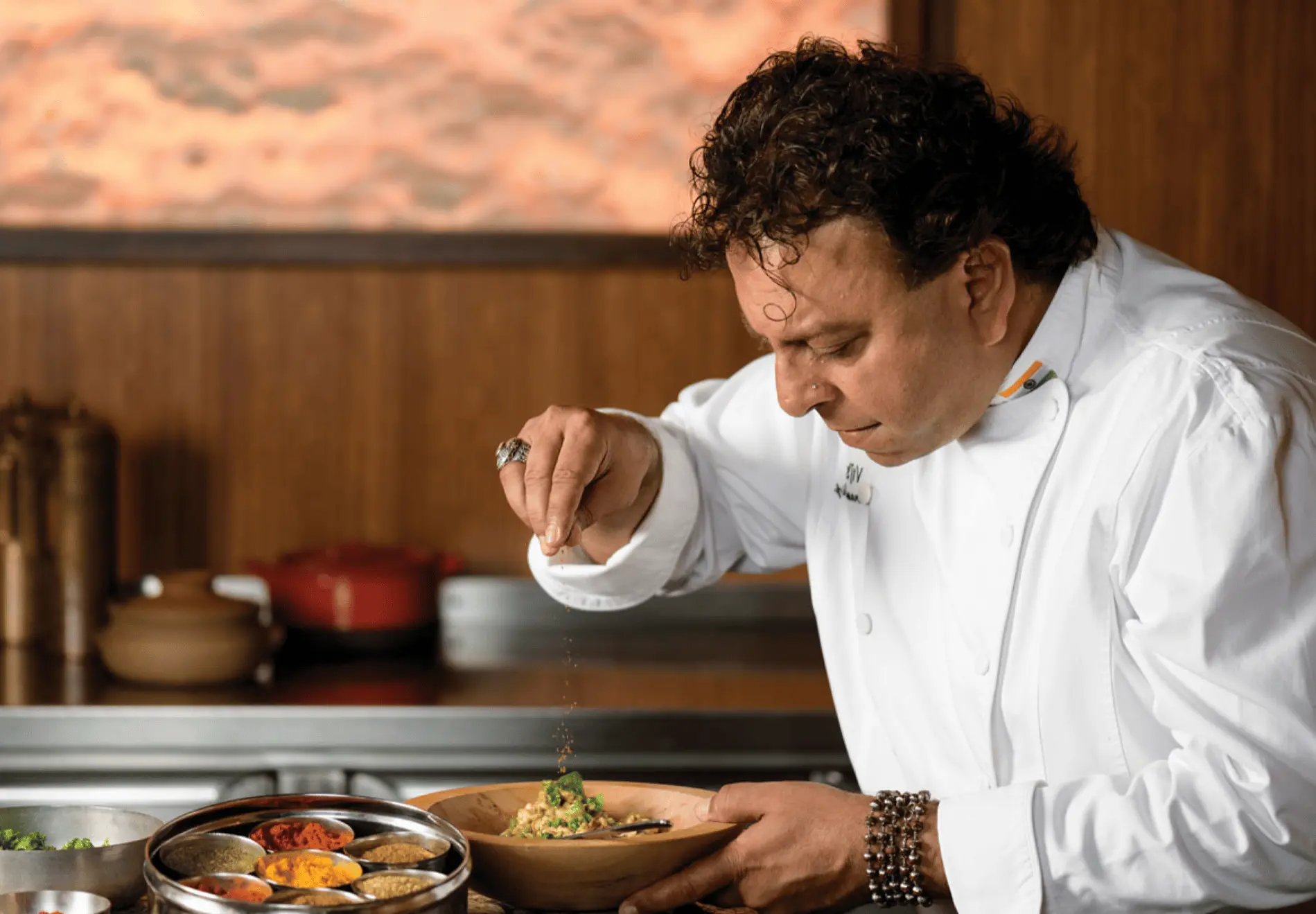 A chef in a white coat sprinkles seasoning onto a dish in a wooden bowl in a kitchen setting.