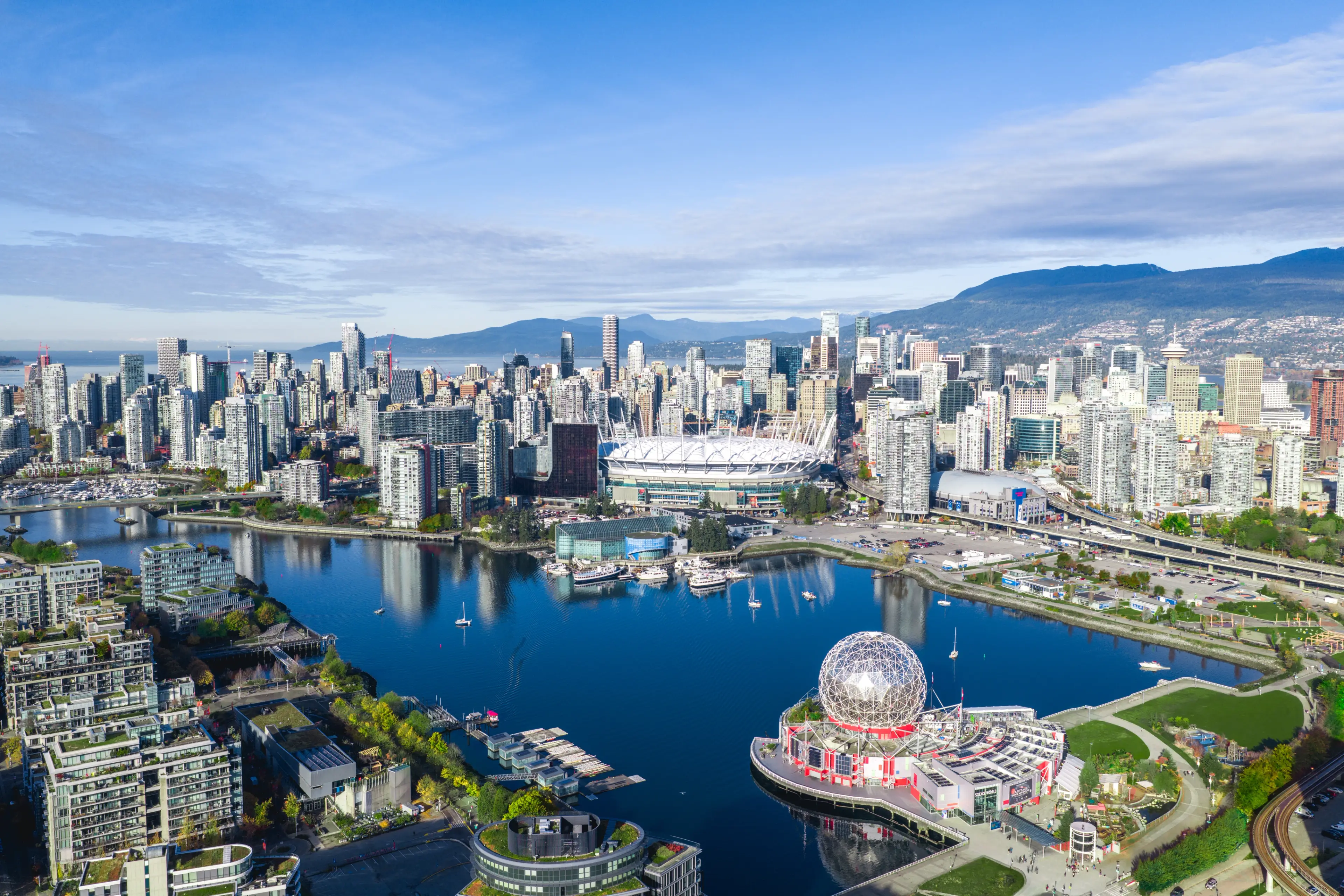 A city skyline with tall buildings, a large stadium, a spherical science museum, and a body of water reflecting the structures under a blue sky.
