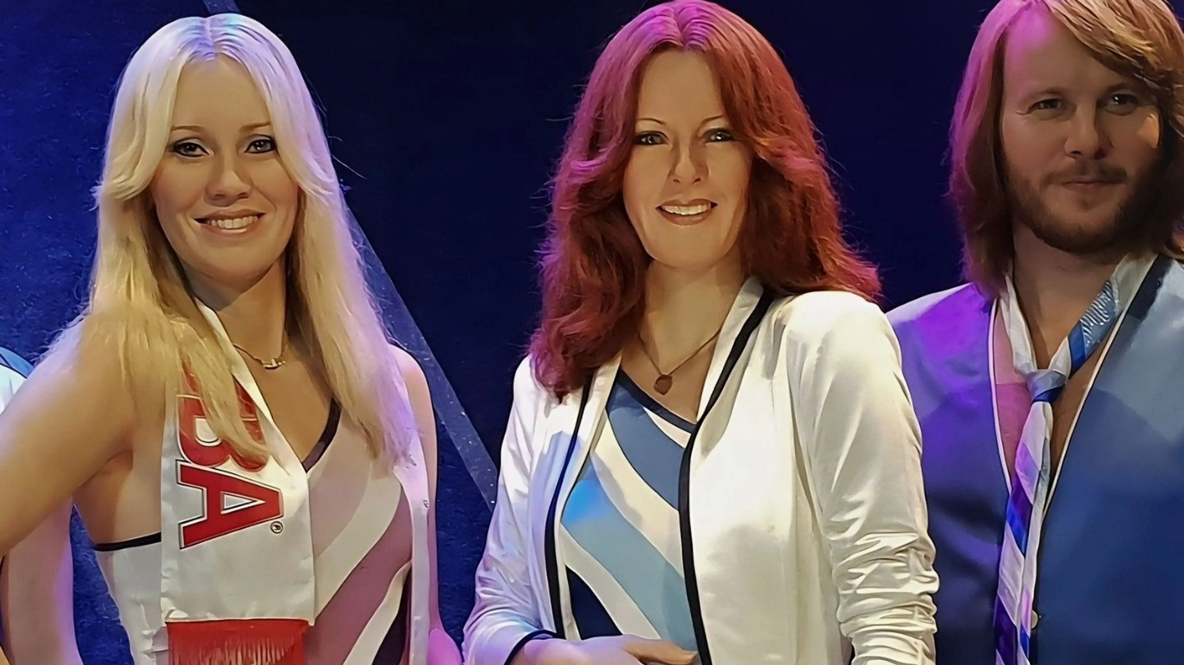 Three people with 1970s style hair and clothing, two with long hair and one with medium-length hair, smiling against a dark background.