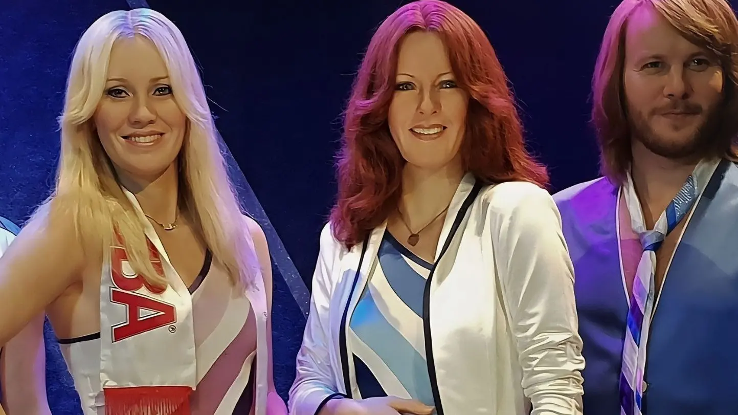 Three people with 1970s style hair and clothing, two with long hair and one with medium-length hair, smiling against a dark background.