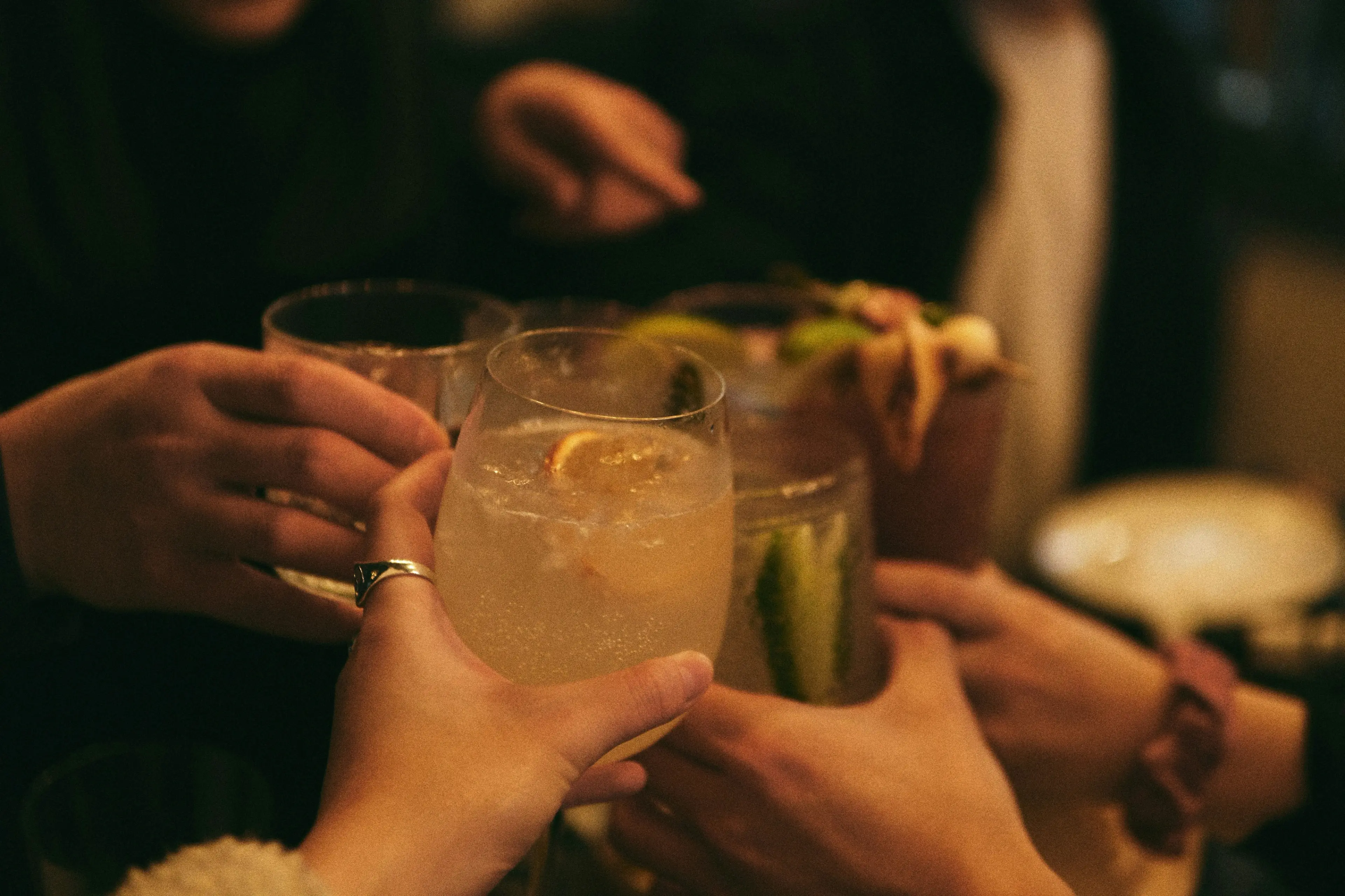 Several hands holding different cocktails and toasting in a dimly lit setting.