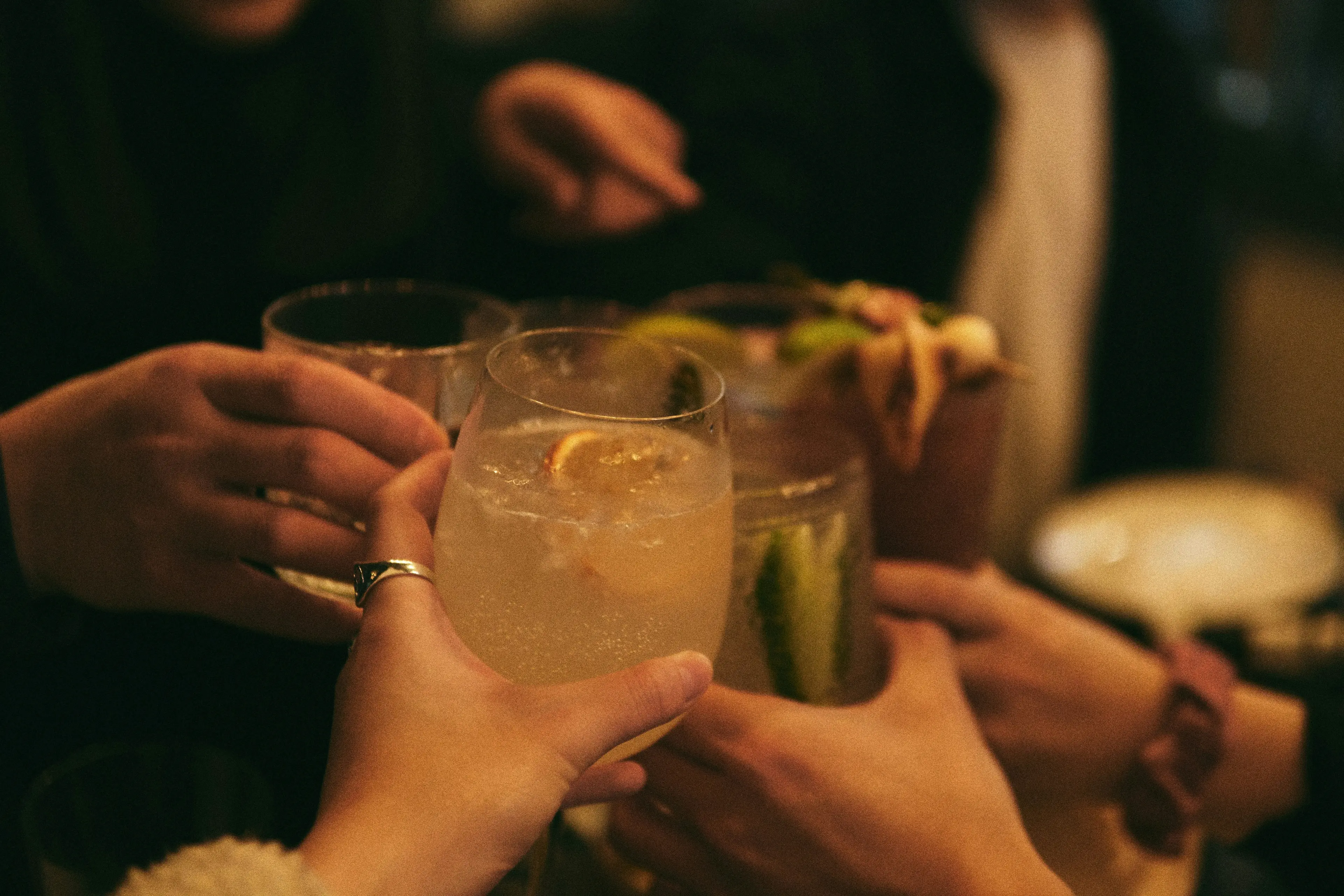 Several hands holding different cocktails and toasting in a dimly lit setting.